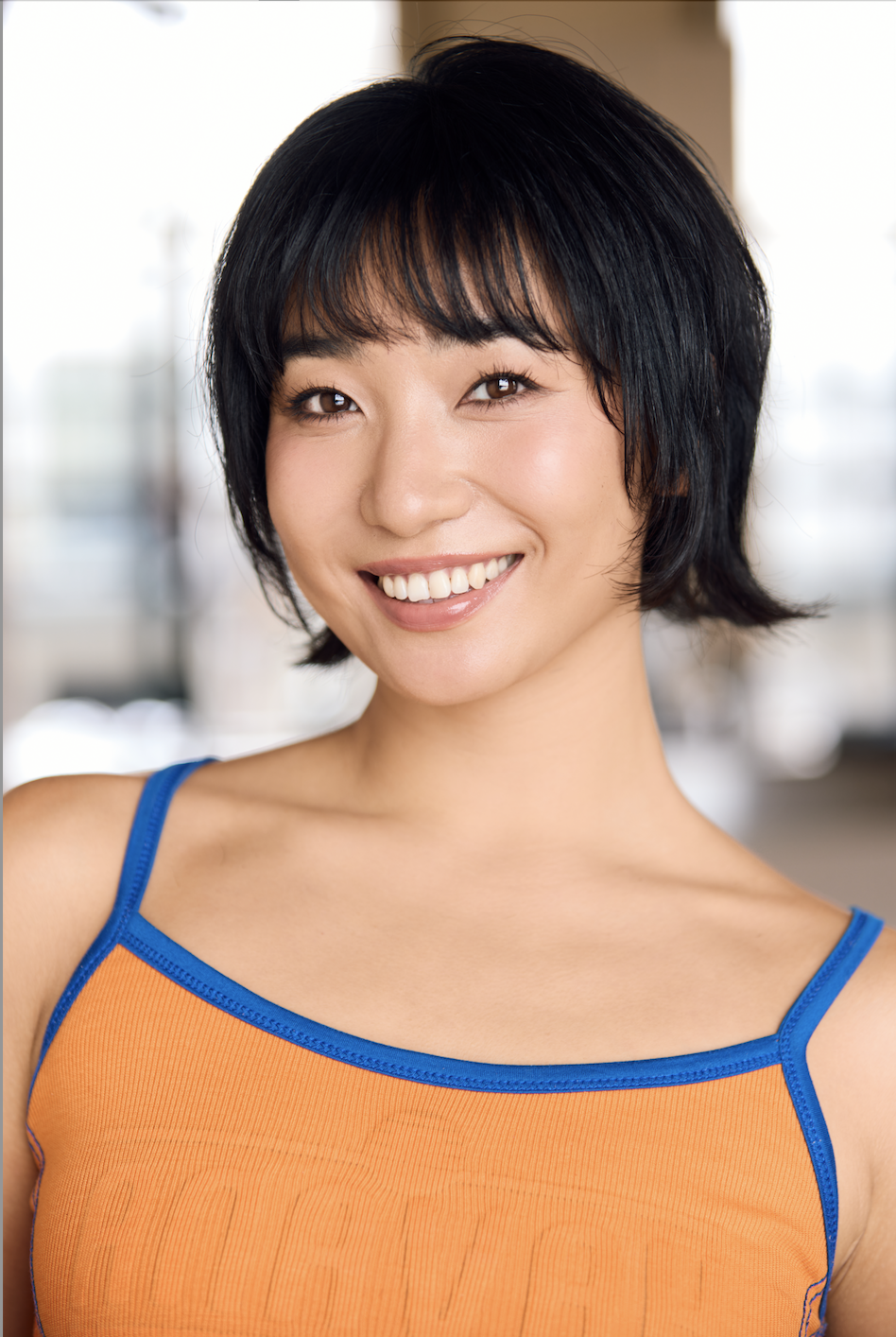 A young woman with short black hair smiling and wearing an orange tank top with blue trim, standing indoors with a blurred background.