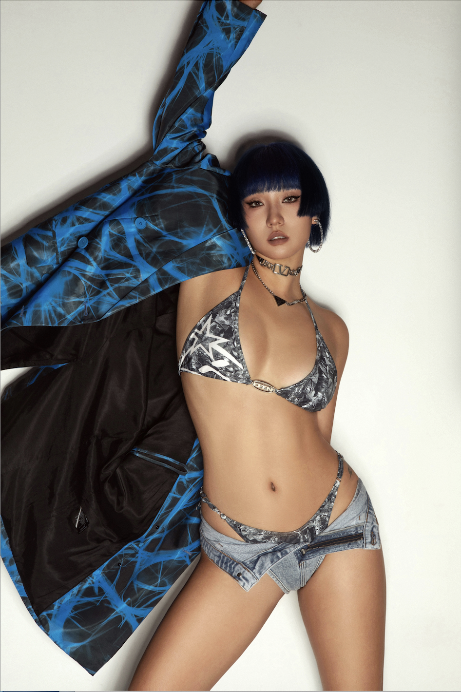 Fashion model with dark blue hair in a bob style wearing a black and white patterned bikini top, denim shorts, and layered gold jewelry, posing against a plain background with her arm raised.