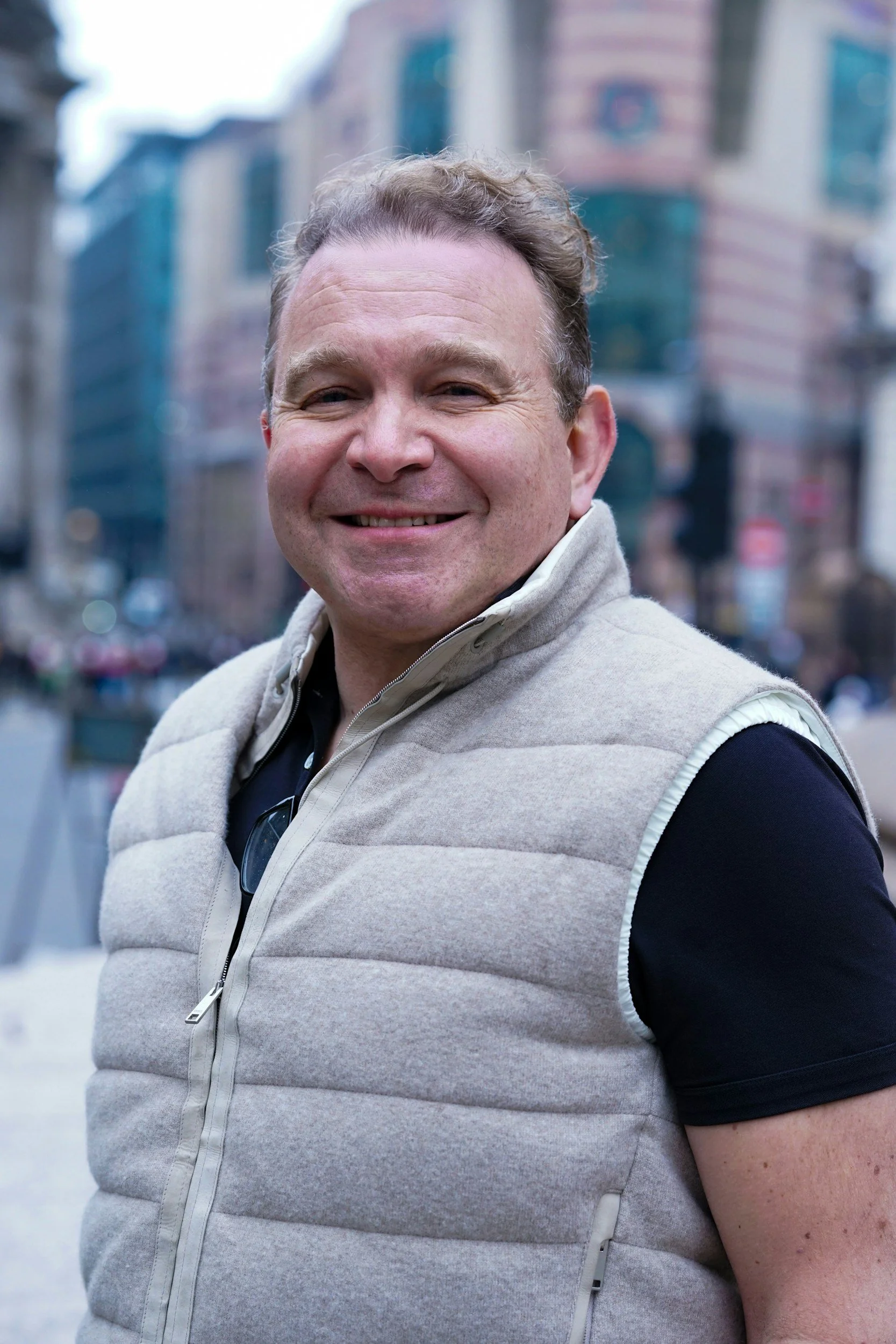 Close-up of a smiling man with curly hair wearing a light-colored vest over a black shirt and sunglasses around his neck, standing outdoors in an urban area with tall buildings in the background.