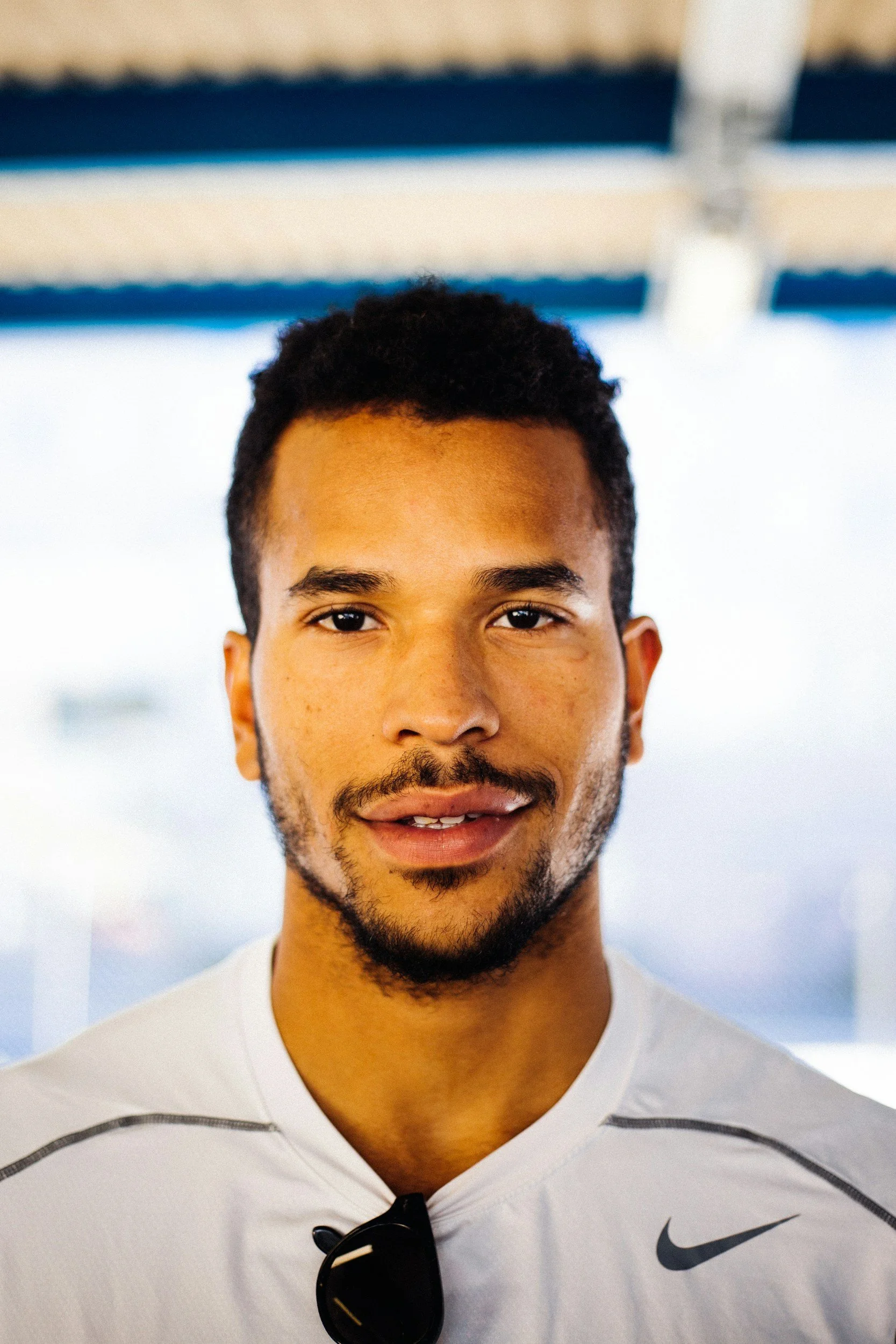 Close-up of a young man's face with short curly hair and a beard, wearing a white Nike sports shirt with black sunglasses hanging on the shirt, standing in an outdoor setting with a blurred background.