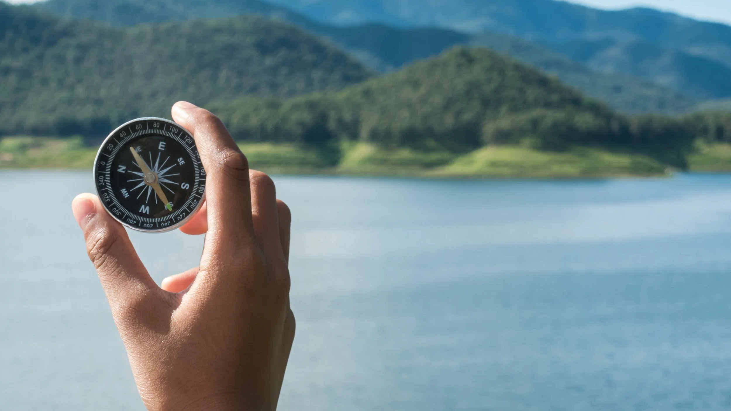 Hand holding a compass with a lake and green mountains in the background.