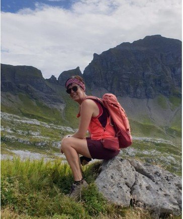 A woman in a pink tank top, purple shorts, and a headscarf crouches on a rock in a mountainous landscape with green grass and rugged peaks in the background.