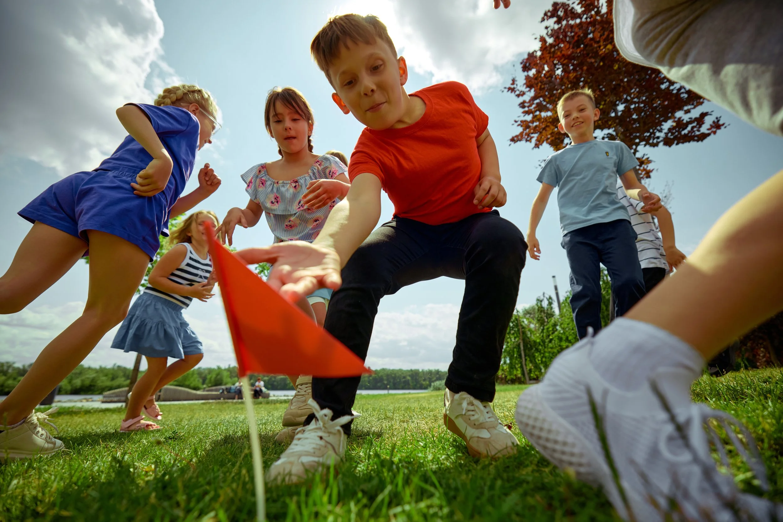 Children playing a game outdoors on a grassy field with a lake and trees in the background, seen from a low angle.
