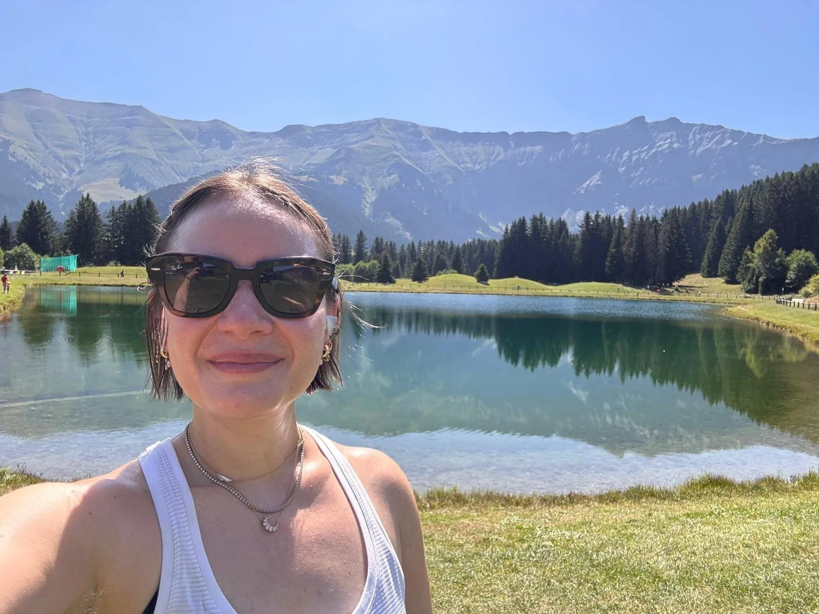 A woman wearing sunglasses taking a selfie near a lake with mountains and trees in the background on a sunny day.