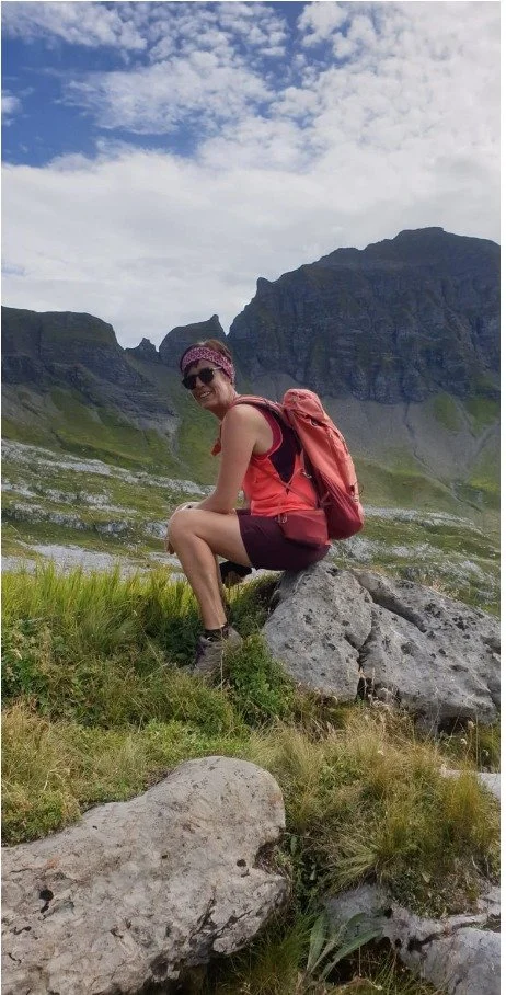 A woman with a pink headband, sunglasses, and a red backpack sitting on a rock in a mountainous area with green grass and cloudy sky.