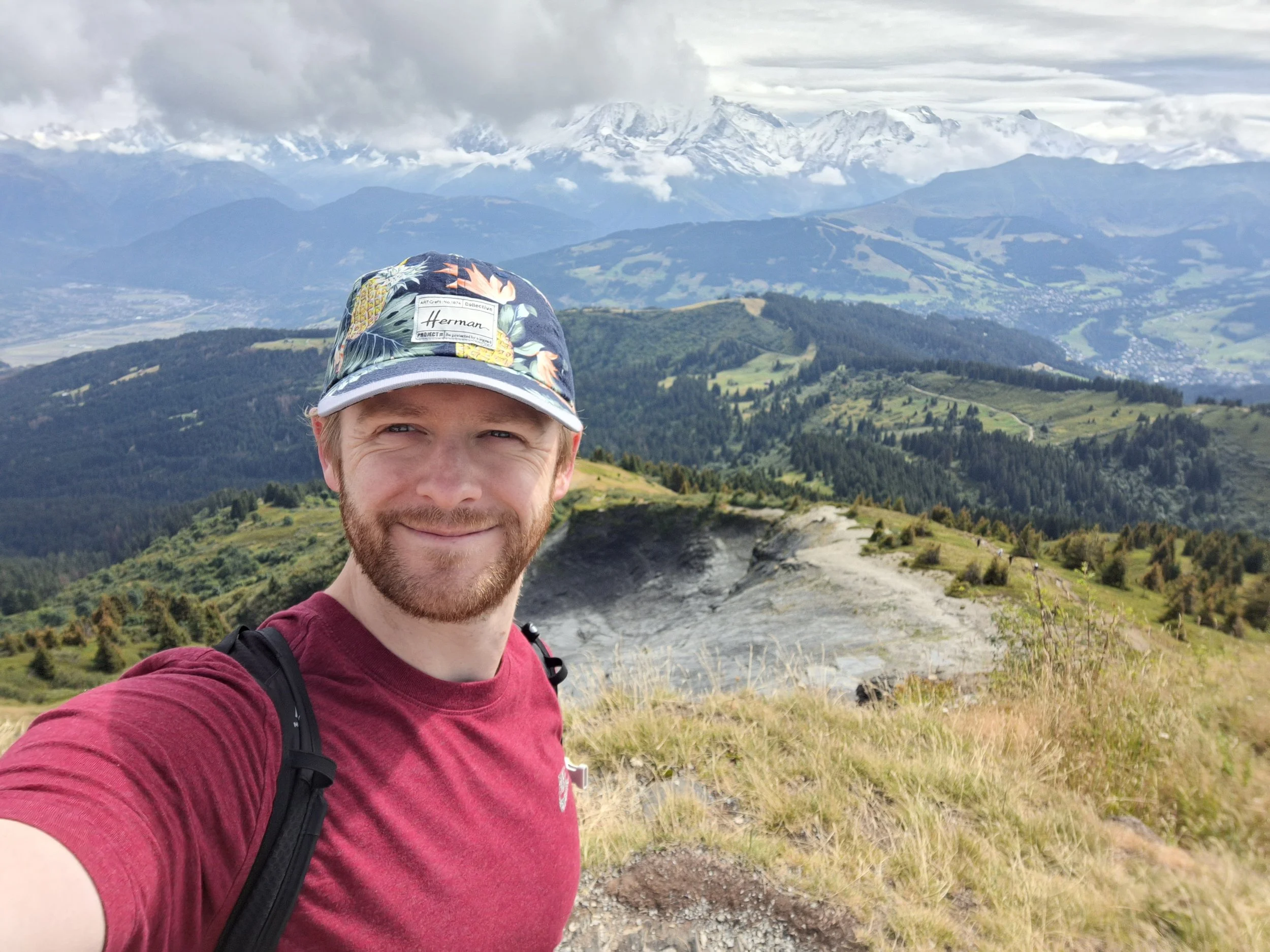 A man with a beard and mustache taking a selfie in a mountainous landscape with snow-capped peaks and green rolling hills in the background. He is wearing a red T-shirt, a backpack, and a cap with a Hawaiian floral pattern.