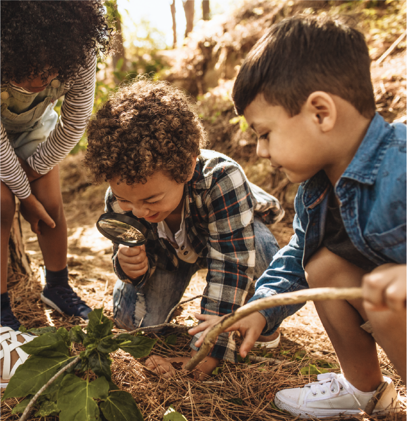 Three children exploring outdoors, with one using a magnifying glass to examine plants and soil on the ground.