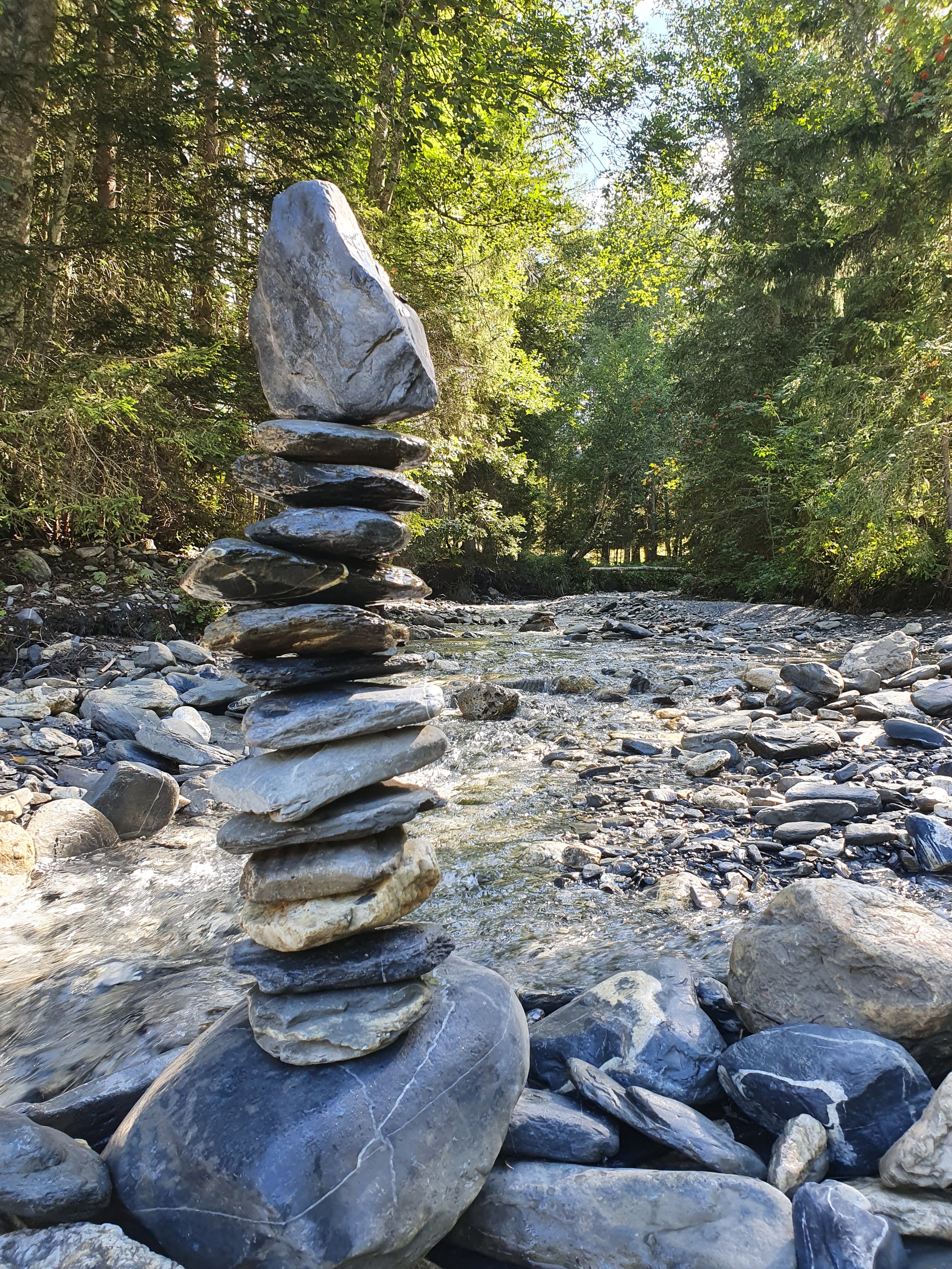 Stacked rocks in a mountain stream with lush green trees and sunlight filtering through the leaves.