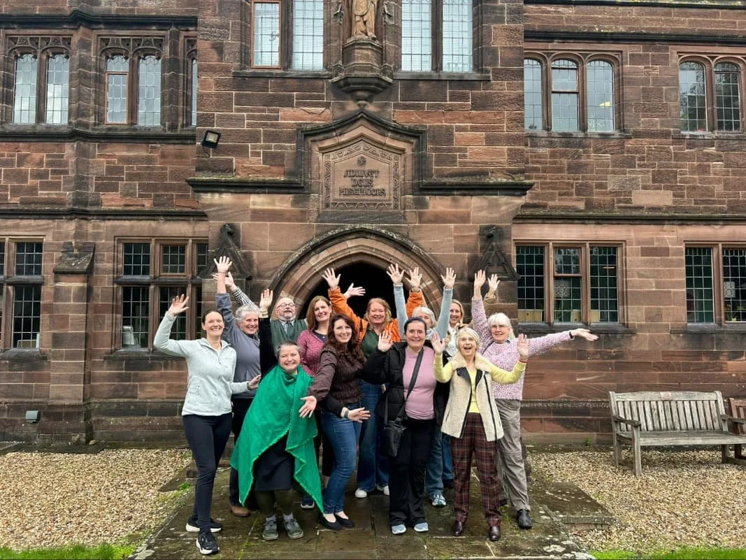 Group of people standing in front of a historic stone building with arched doorway, some raising their hands and smiling.