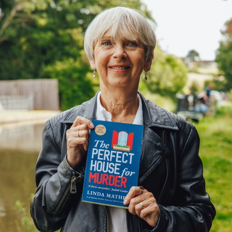 Linda Mather wearing a leather jacket holding her latest book "the perfect house for murder" with the canal in the background.