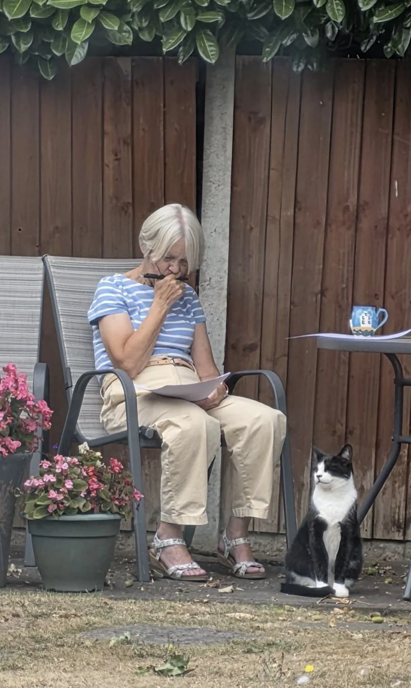 An elderly woman with white hair, wearing a blue and white striped shirt and beige pants, sitting outside on a patio. She is holding a pen and some papers, possibly reading or writing, with a black and white cat sitting nearby on the ground. There are pink flowers in pots beside her and a mug on a table nearby, all against a wooden fence and leafy plants overhead.