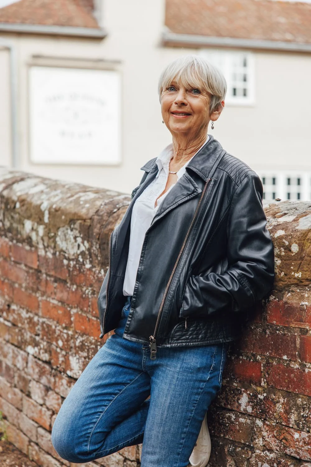 Linda Mather wearing a leather jacket and jeans stood against a red brick wall smiling at the camera.