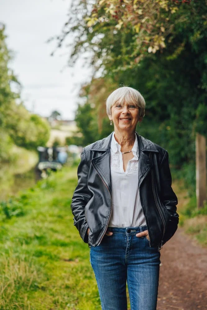 Linda smiling and walking outdoors on a trail, wearing a black leather jacket, white blouse, and blue jeans, with a lush green background.