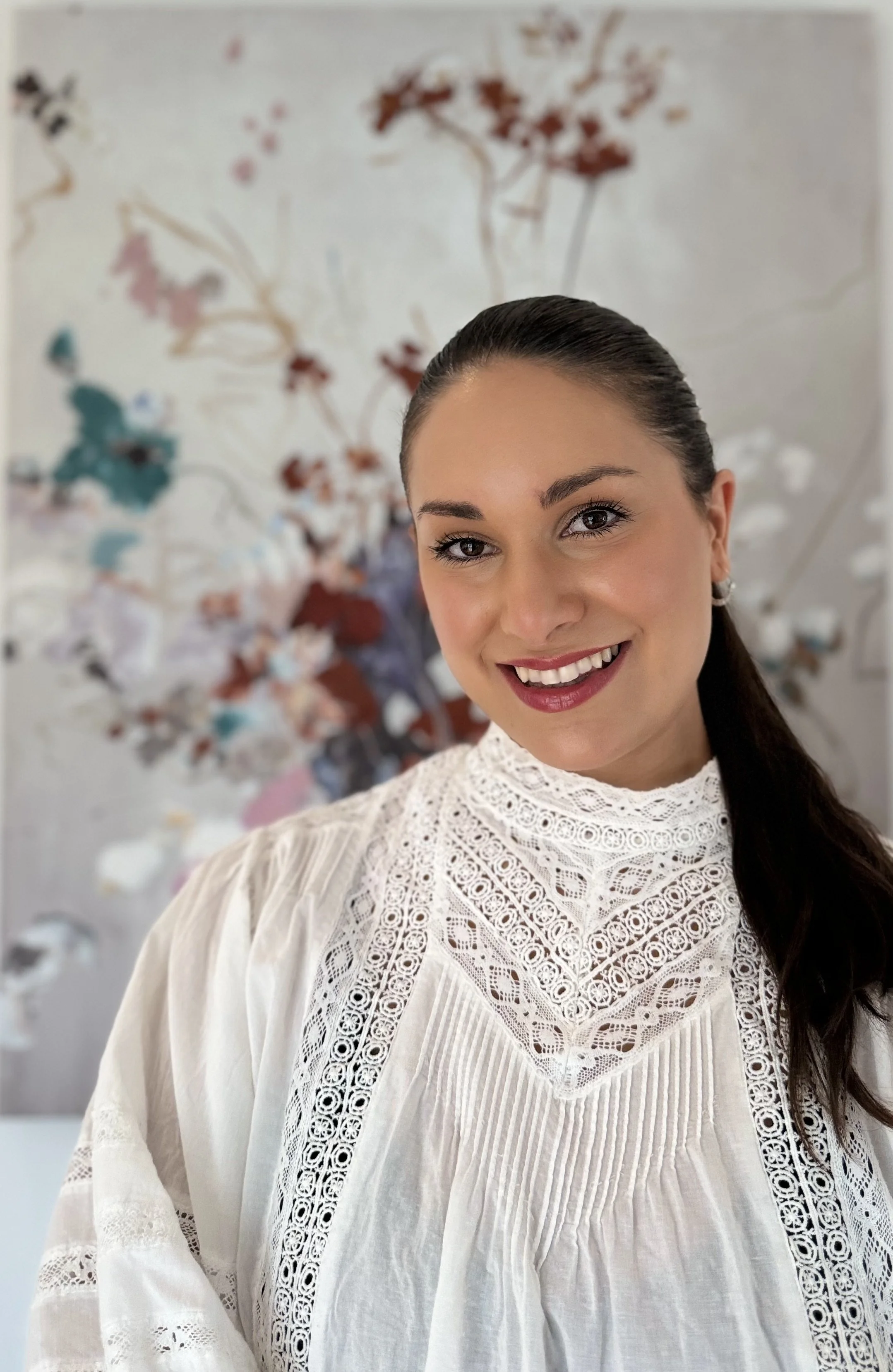 Et bilde av en smilende kvinne med mørkt hår som har brukt en hvit, broderet alpakkakleid. Hun står foran et abstrakt kunstverk med blomsterliknende mønstre i ulike farger.