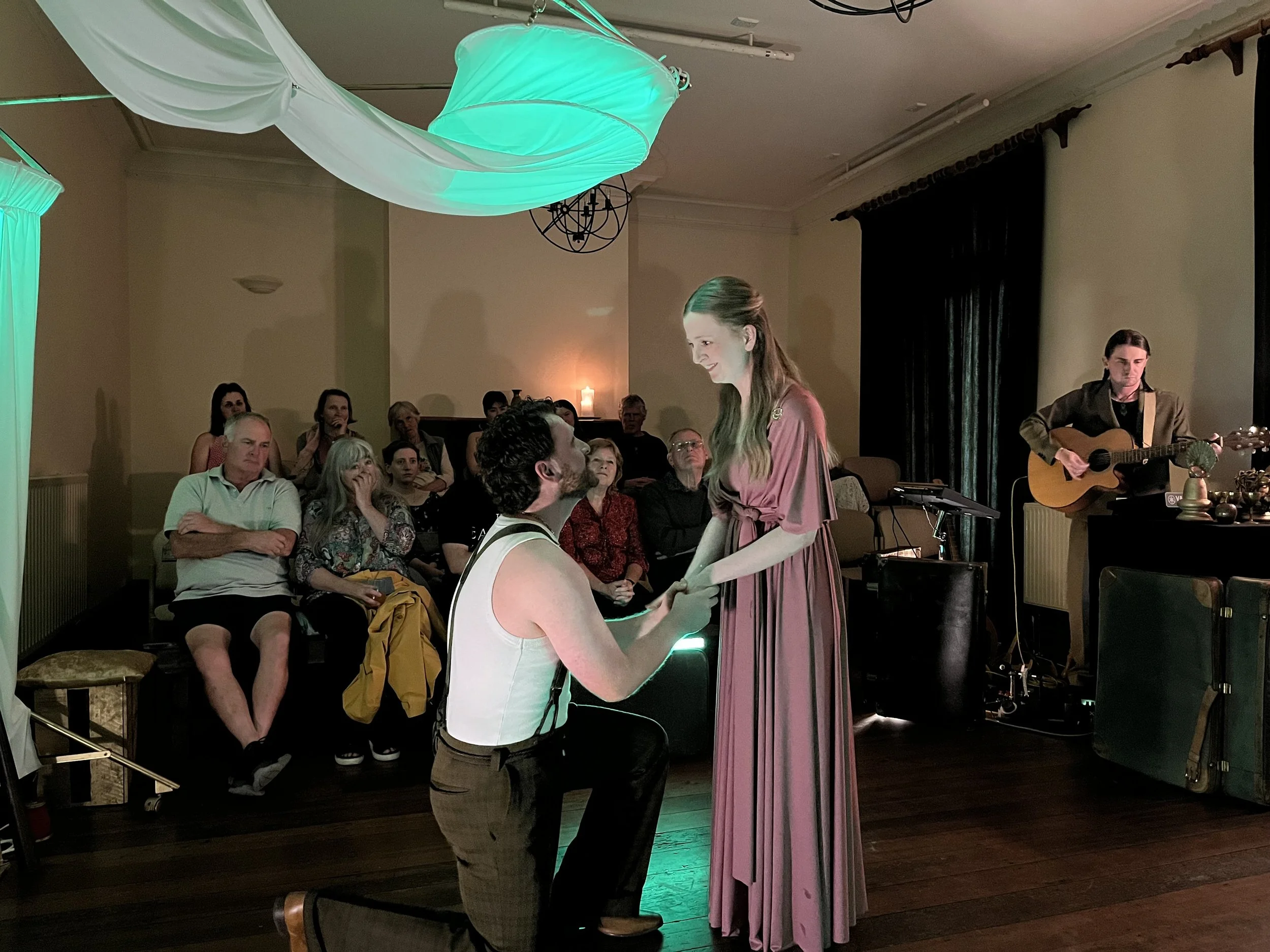 A woman in a pink dress is standing and smiling at a man sitting on a chair, with a guitarist playing nearby, in a cozy indoor setting with an audience watching them perform.
