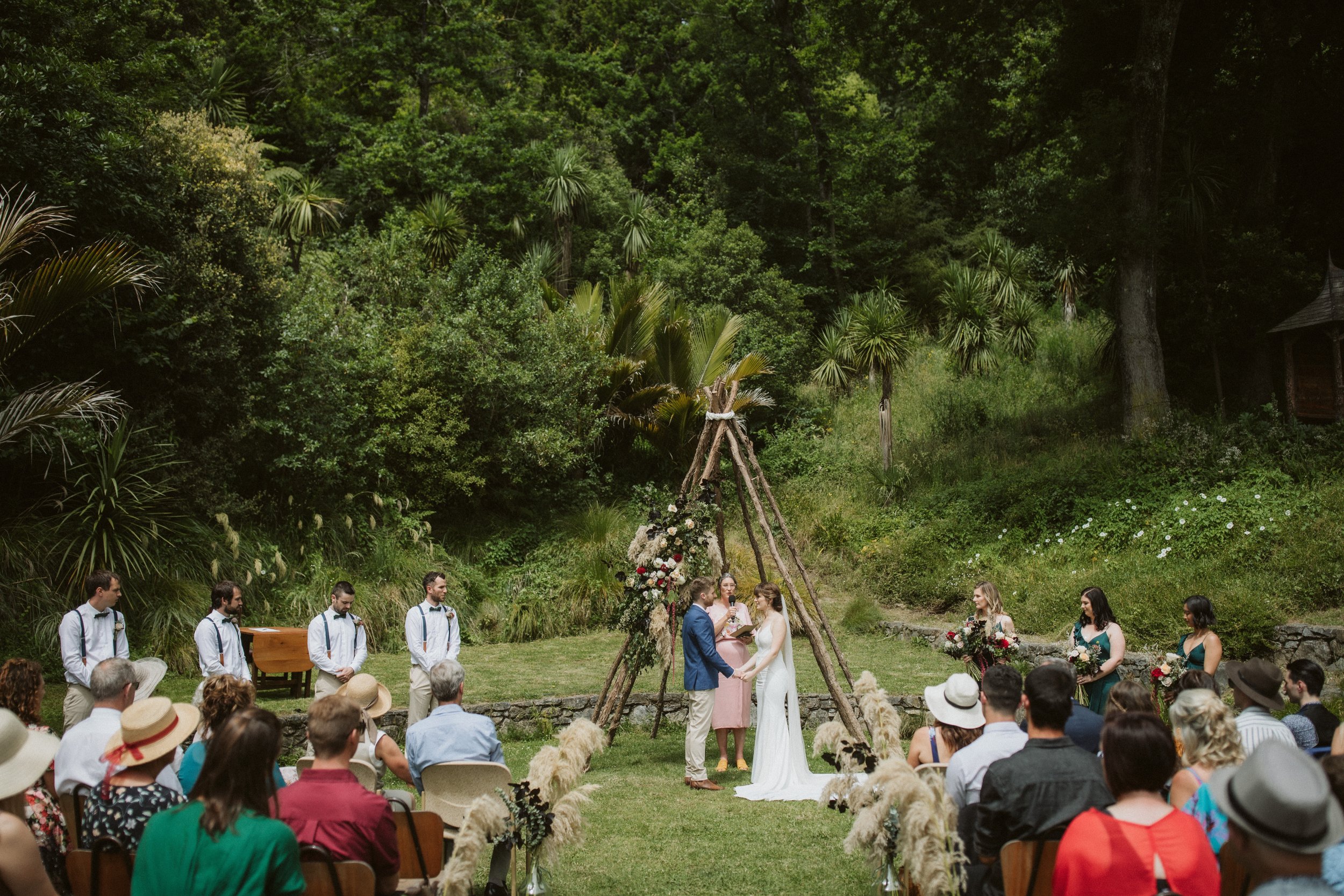 Outdoor wedding ceremony taking place in a lush green forest, with a bride and groom exchanging vows under a wooden arch decorated with flowers, surrounded by guests seated and standing, some wearing hats.