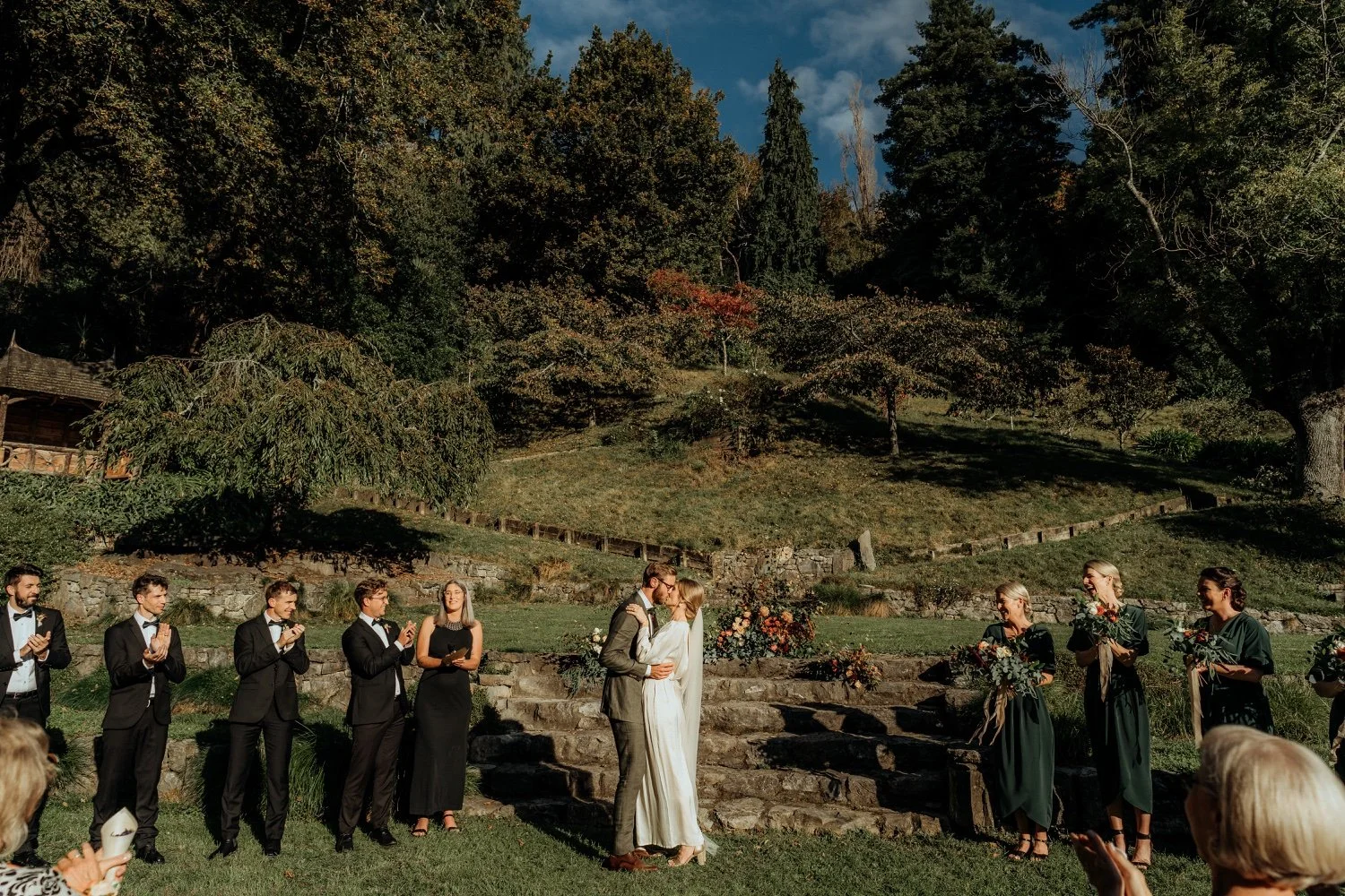 A wedding ceremony outdoors with the bride and groom kissing on stone steps, surrounded by bridesmaids holding bouquets and groomsmen in suits, against a backdrop of trees and blue sky.
