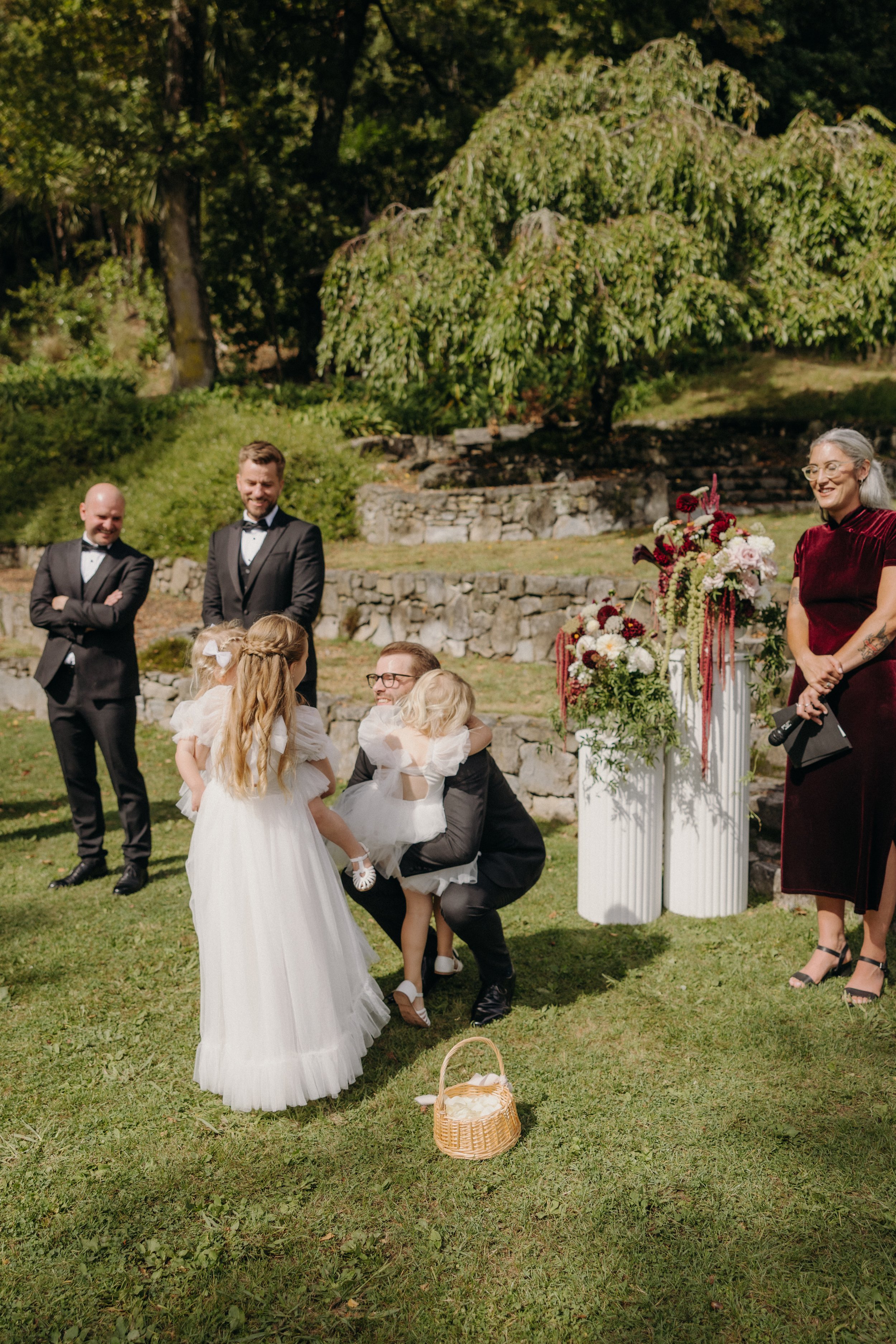 Children dressed in white dresses and black suits gather outdoors on a grassy area at a wedding ceremony, surrounded by adults in formal attire, with decorative flowers on white columns and trees in the background.