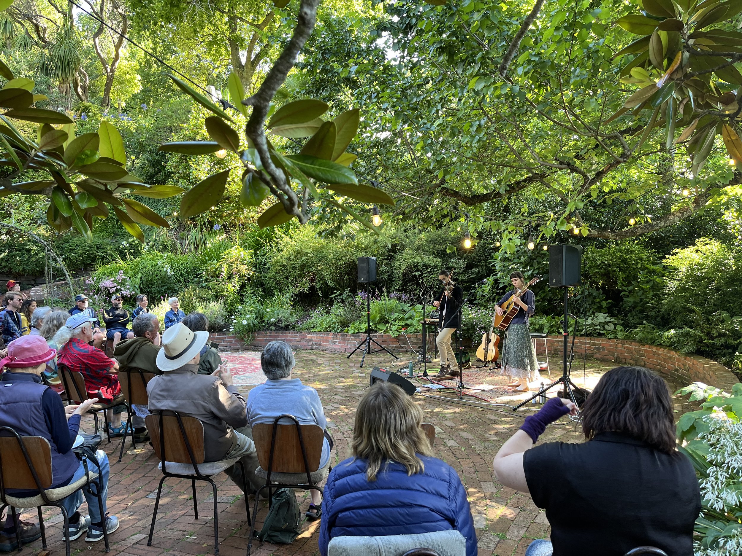 A group of people attending an outdoor live music concert in a lush green garden with trees and hanging string lights. The band is playing on a small brick stage with musical instruments, including guitars.