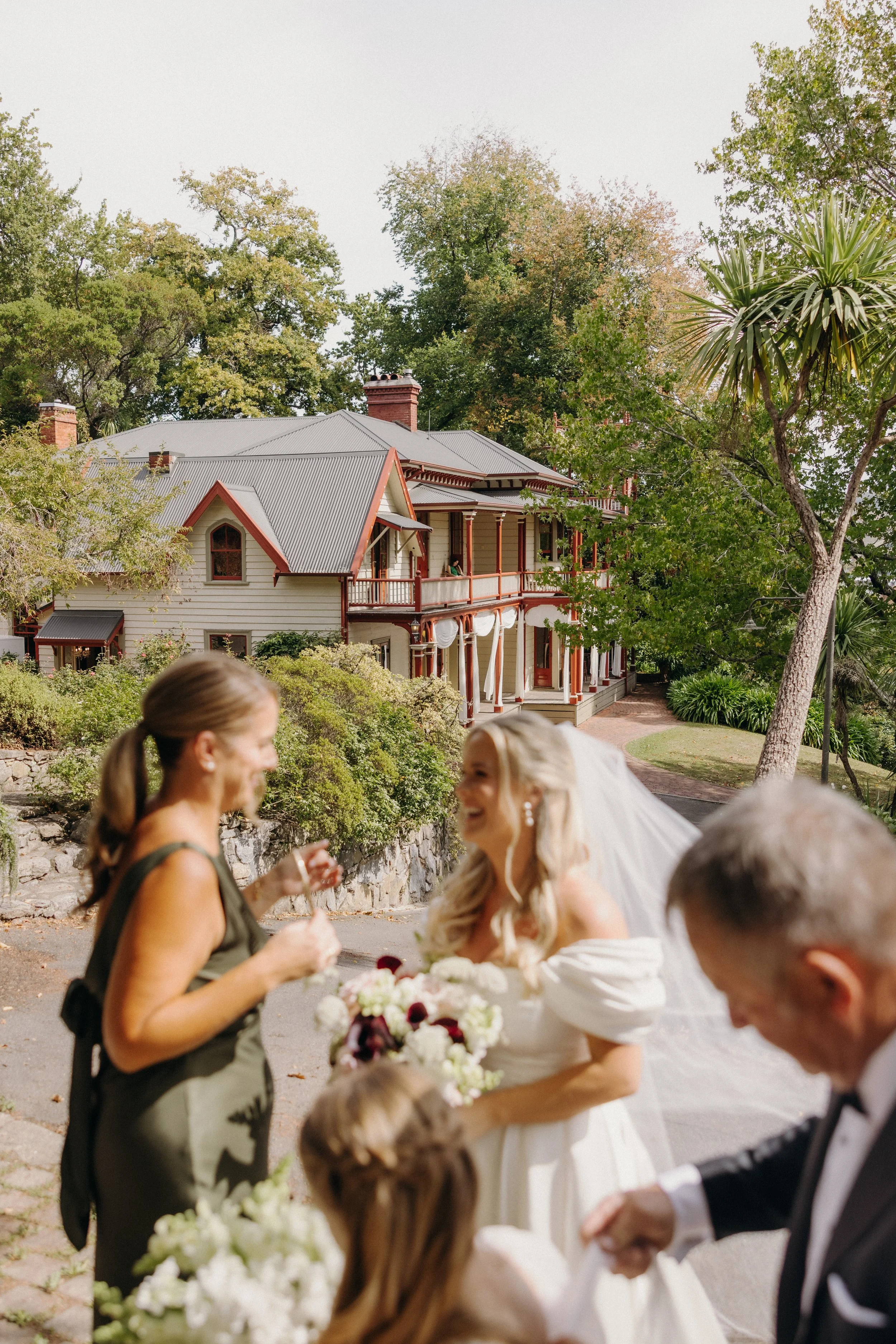 A bride in a white wedding dress holding a bouquet of flowers, smiling and talking with a woman in a green dress at an outdoor wedding reception, with a large house and trees in the background.