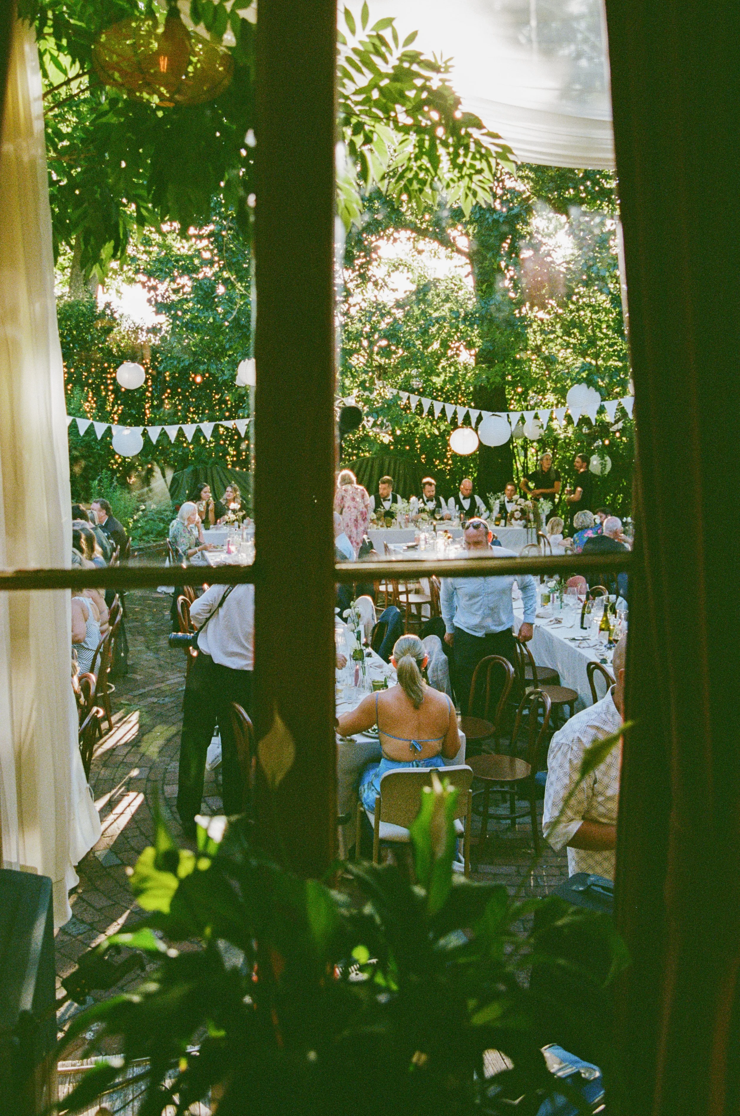 A view through a window showing an outdoor celebration with people dining at tables, festive banners, and string lights hanging among trees during sunset.