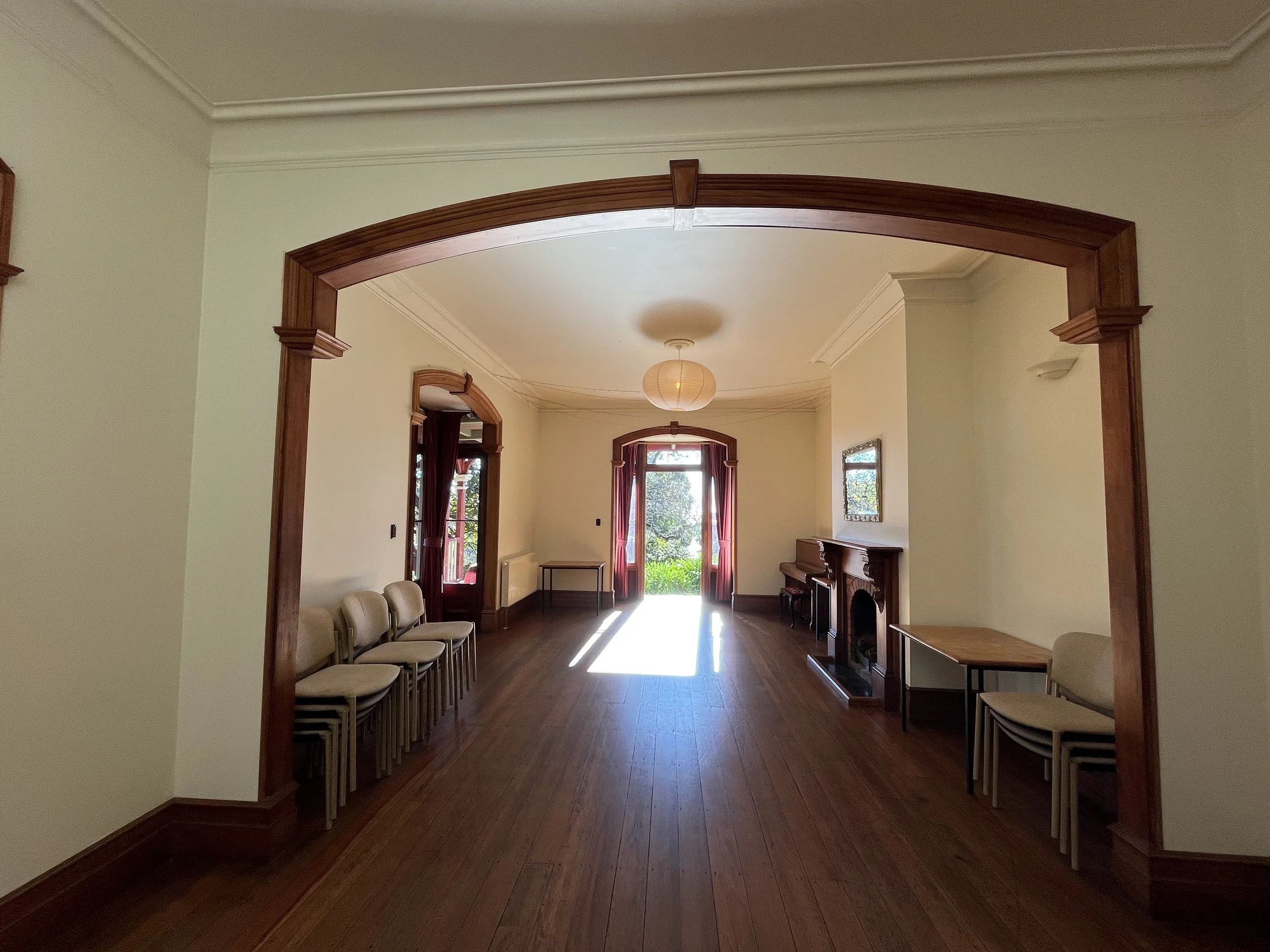 An interior room with wooden flooring, cream-colored walls, and large windows with red curtains. There are chairs lined up against the wall on the left and a fireplace on the right, with a wooden table and a piano near the fireplace. A doorway with a