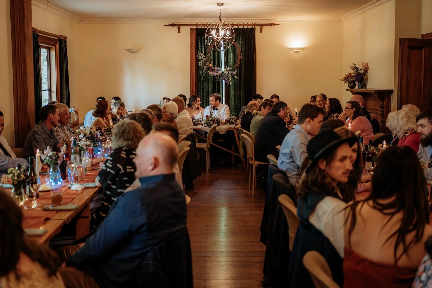 People sitting at a decorated long dining table during a wedding reception or celebration in a warmly lit room with wood trim and green curtains.