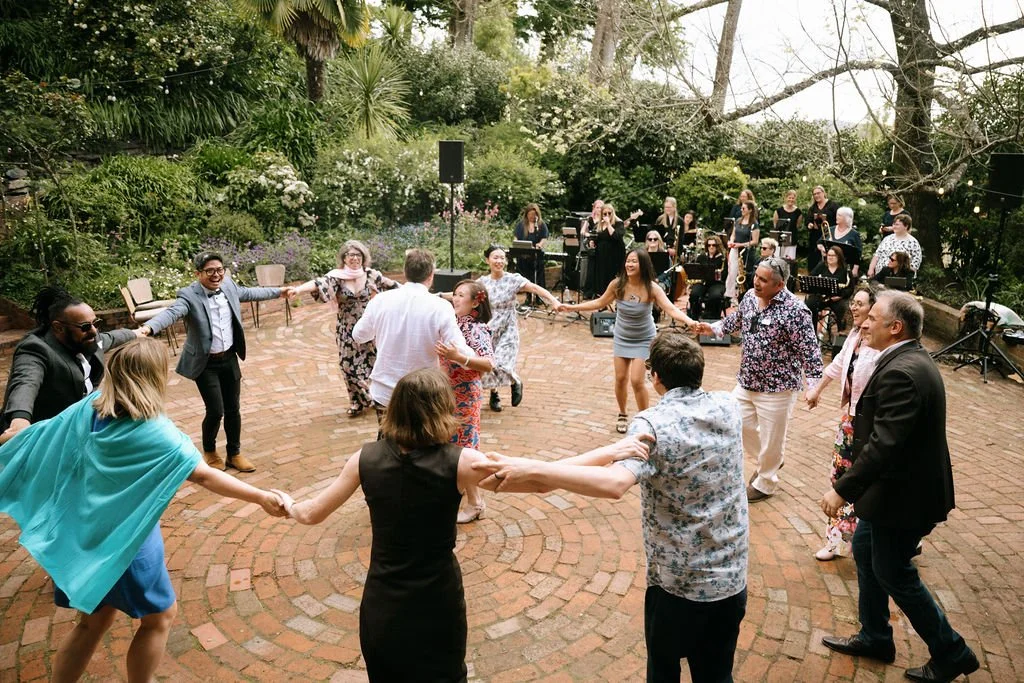 People dancing in a circle at an outdoor party with a live band and lush greenery in the background.
