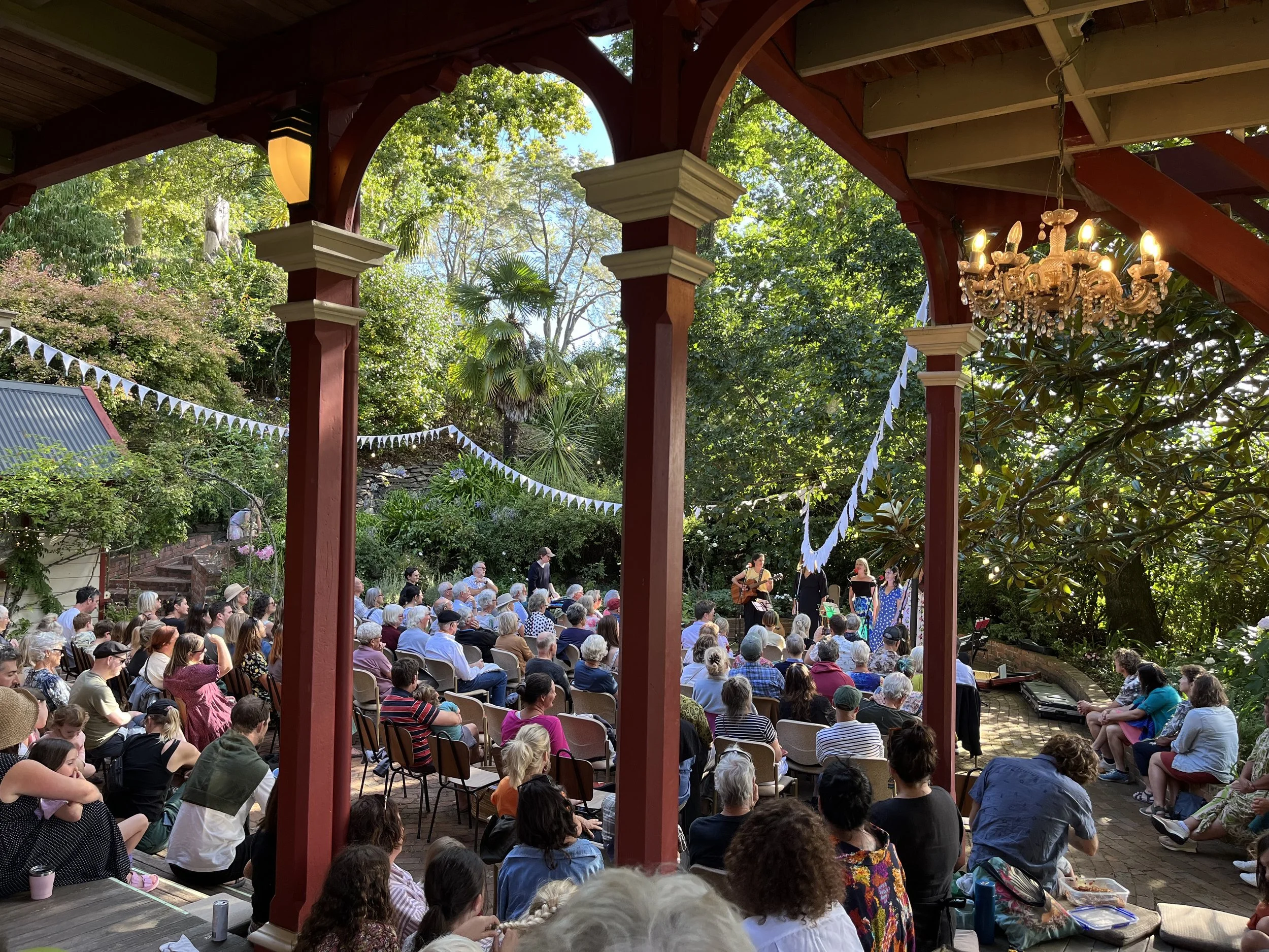 An outdoor concert with a large audience seated in chairs, performing on stage with musicians and singers, surrounded by lush green trees and decorated with white bunting flags, under a red wooden pavilion with a chandelier.
