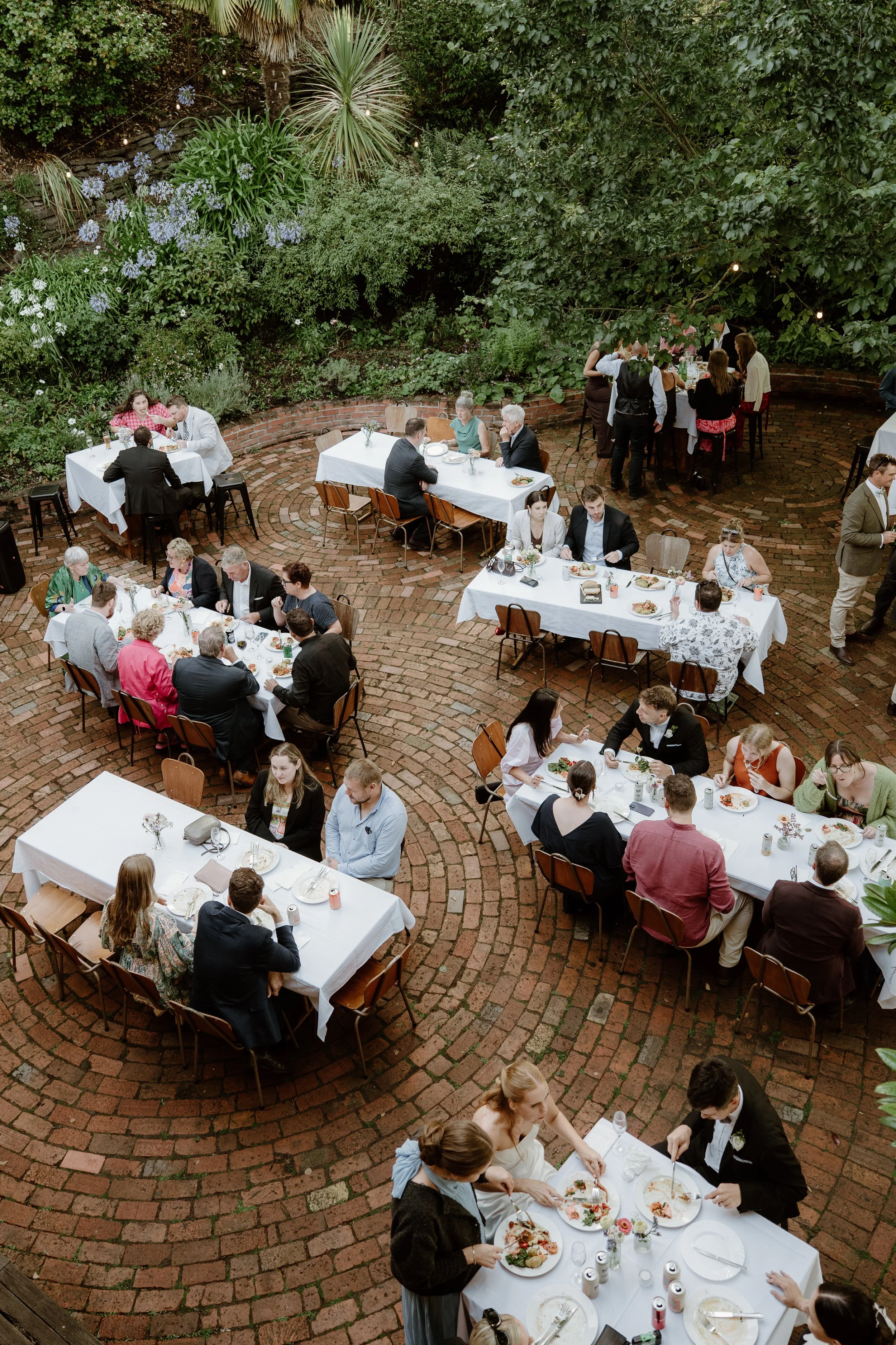 Outdoor social gathering with people seated at tables with white tablecloths, eating and chatting, in a garden setting with trees and string lights.