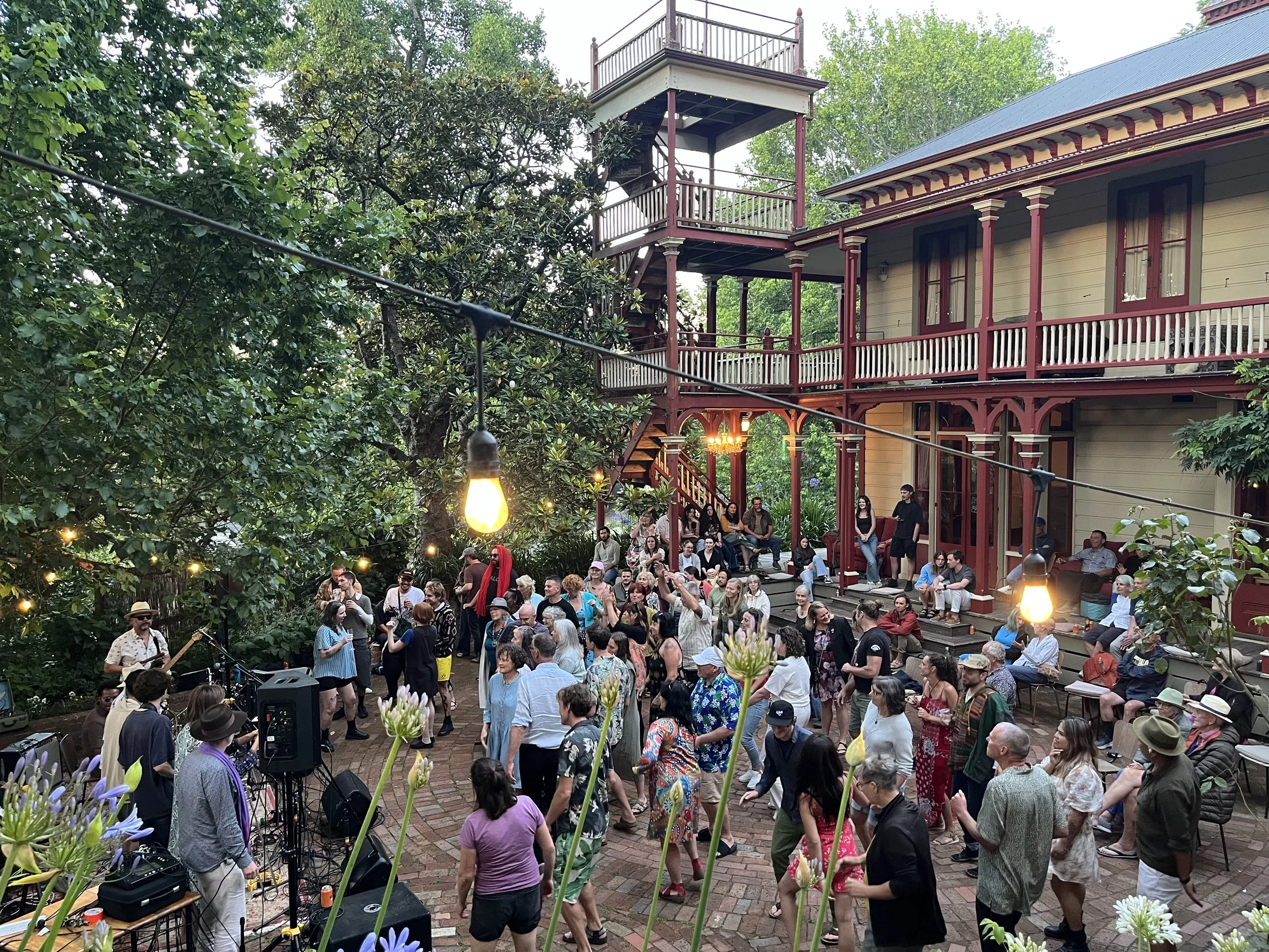 A large group of people gathered outdoors in a garden courtyard, attending a live music event with musicians performing, and an old Victorian-style house with a wrap-around porch in the background.