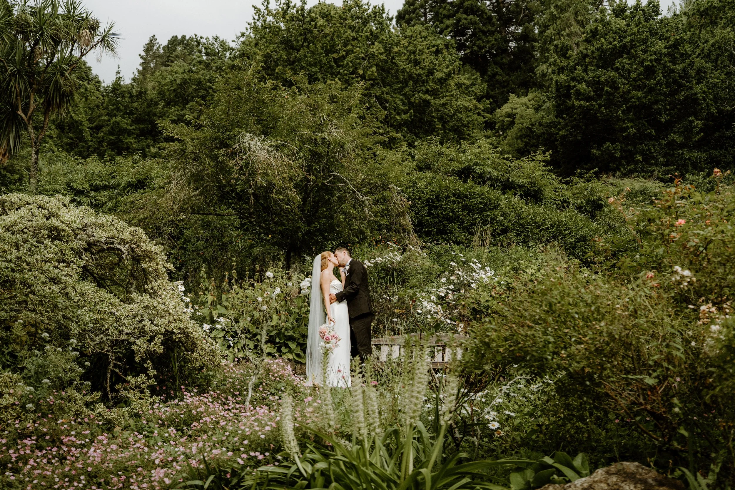 A bride and groom kissing in a lush garden filled with various green plants, flowers, and trees, during a wedding ceremony.