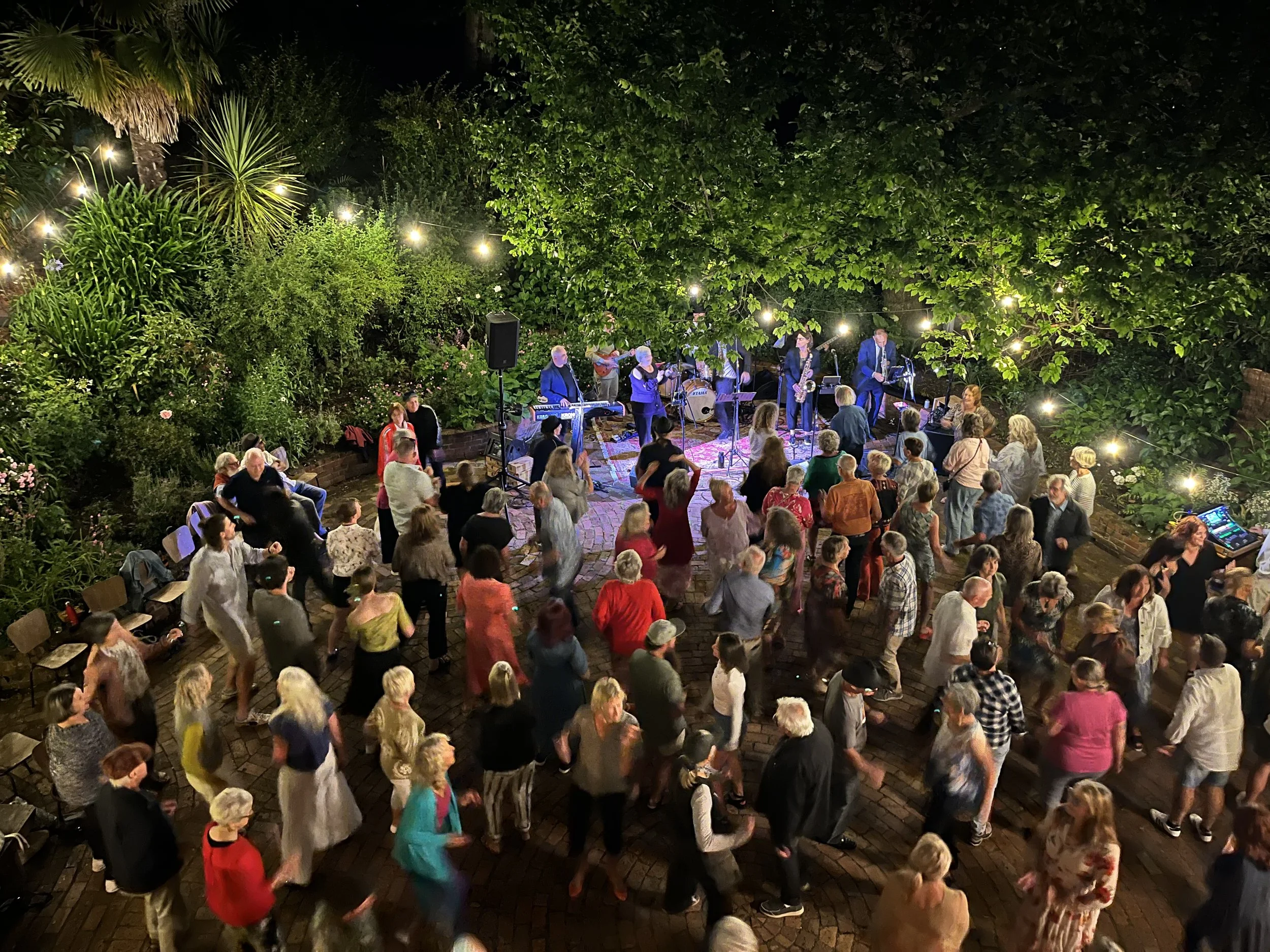 An outdoor night event with a band performing on stage and an audience dancing and sitting in a garden with string lights. Trees and lush greenery surround the area.