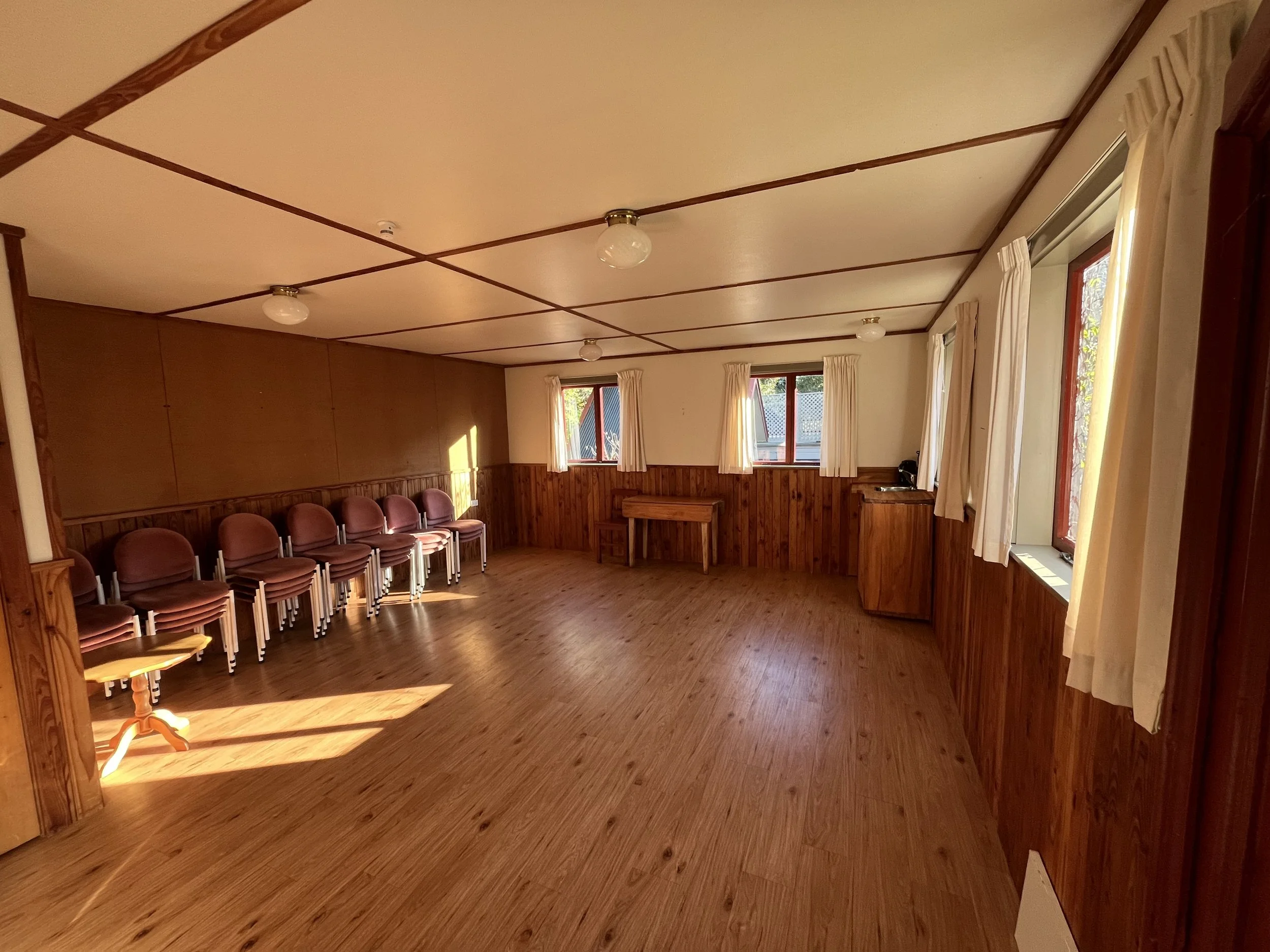 An empty room with wooden walls and ceiling, several set of chairs arranged along the walls, and windows with white curtains letting in sunlight.