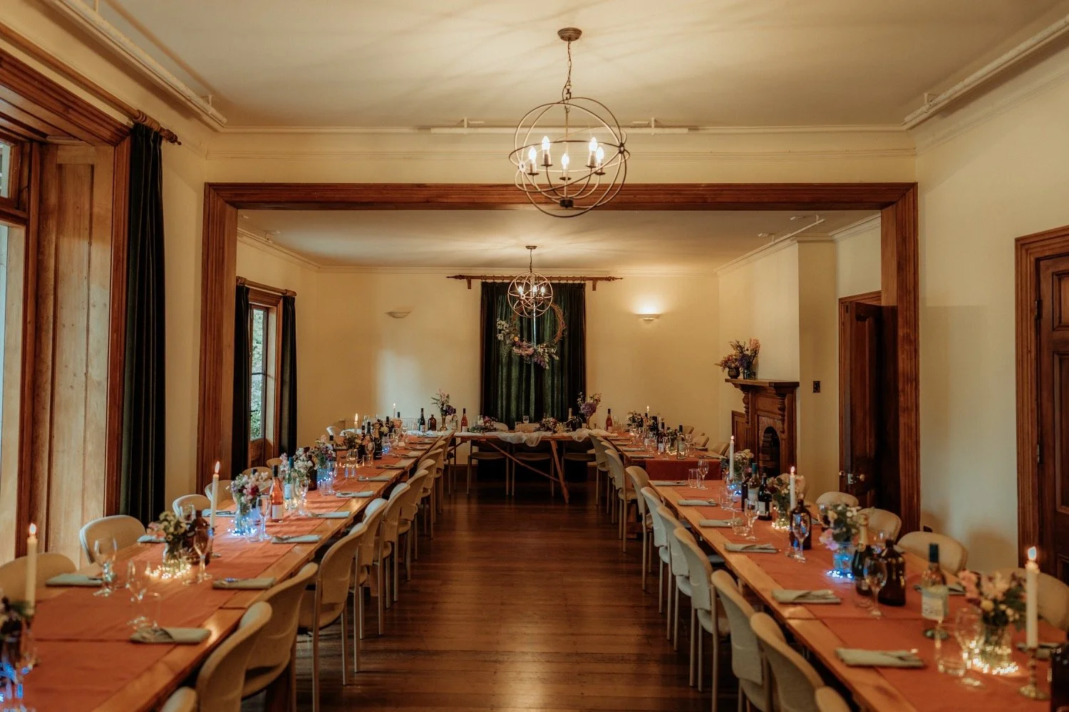 A banquet room decorated for a celebration with long tables covered in peach-colored tablecloths, floral centerpieces, candles, place settings, and wine bottles. The room has wood trim, a fireplace, and chandeliers hanging from the ceiling.