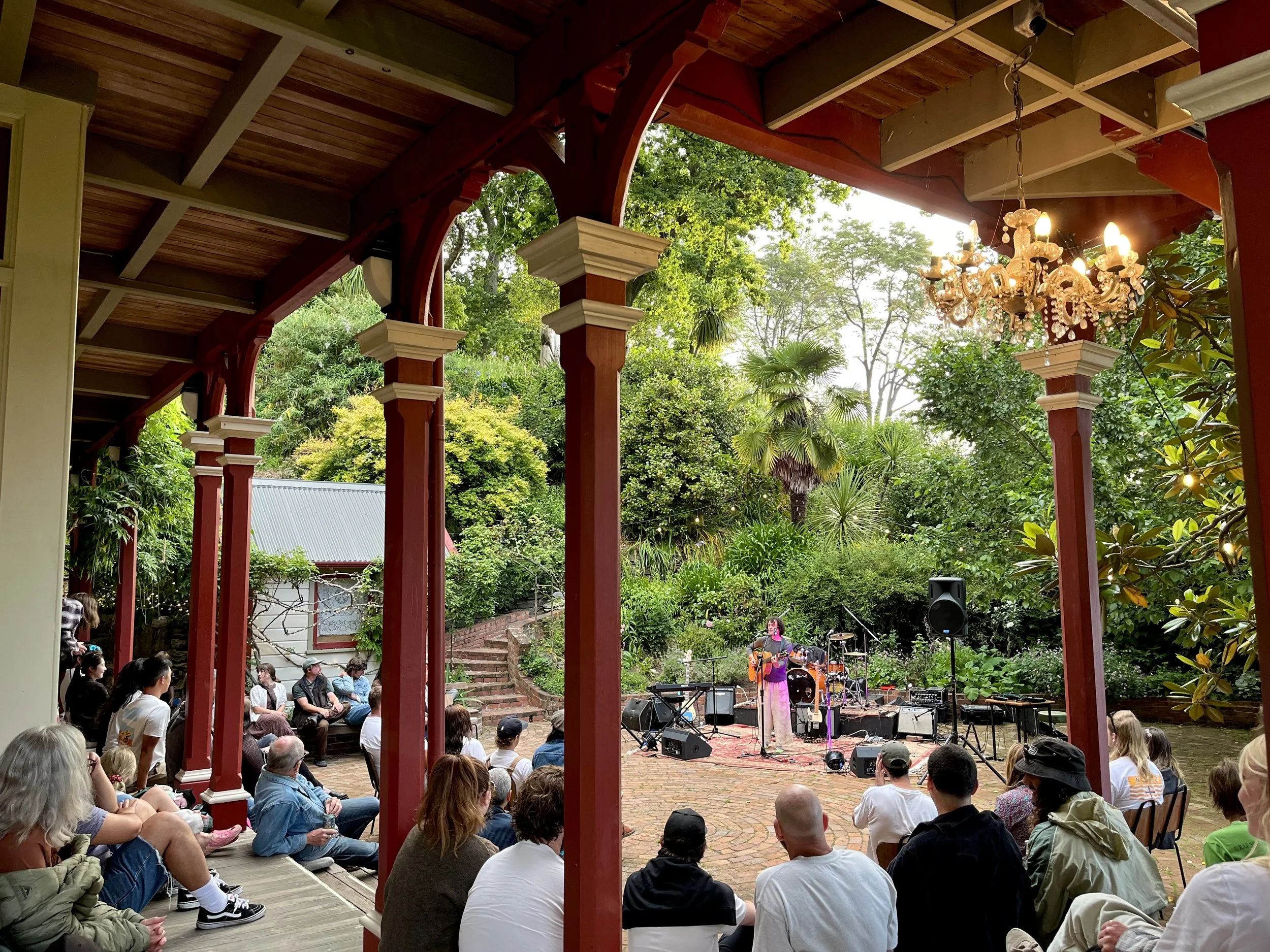 People sitting outdoors watching a live musical performance with a singer and a band, surrounded by lush green trees, under a red wooden pergola, with a chandelier hanging from above.