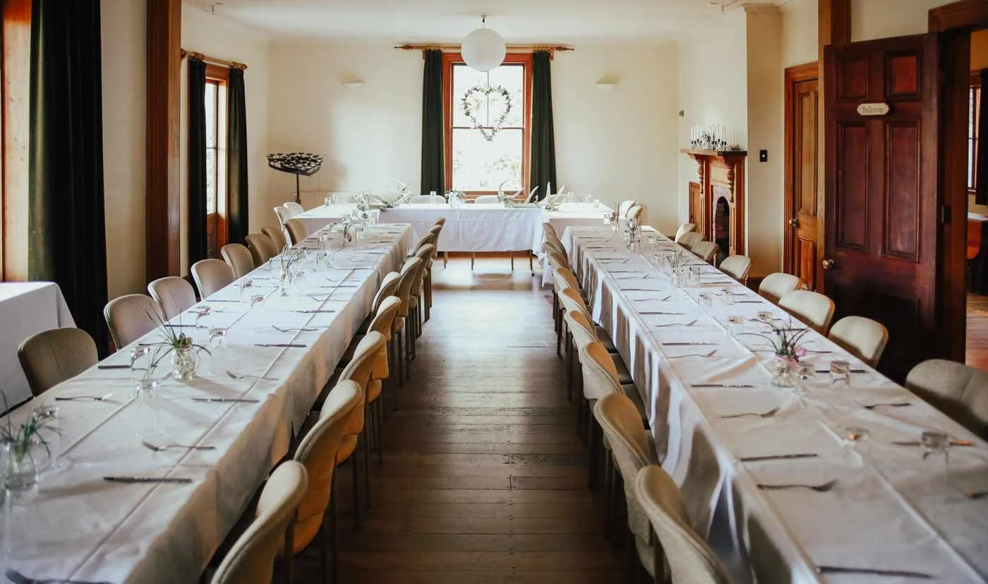 A cozy dining room decorated for a special event with long tables covered in white tablecloths, set with glasses, plates, and silverware, and surrounded by beige chairs. There are windows with dark curtains, a decorative wreath hanging on the middle window, and a fireplace on the right side.