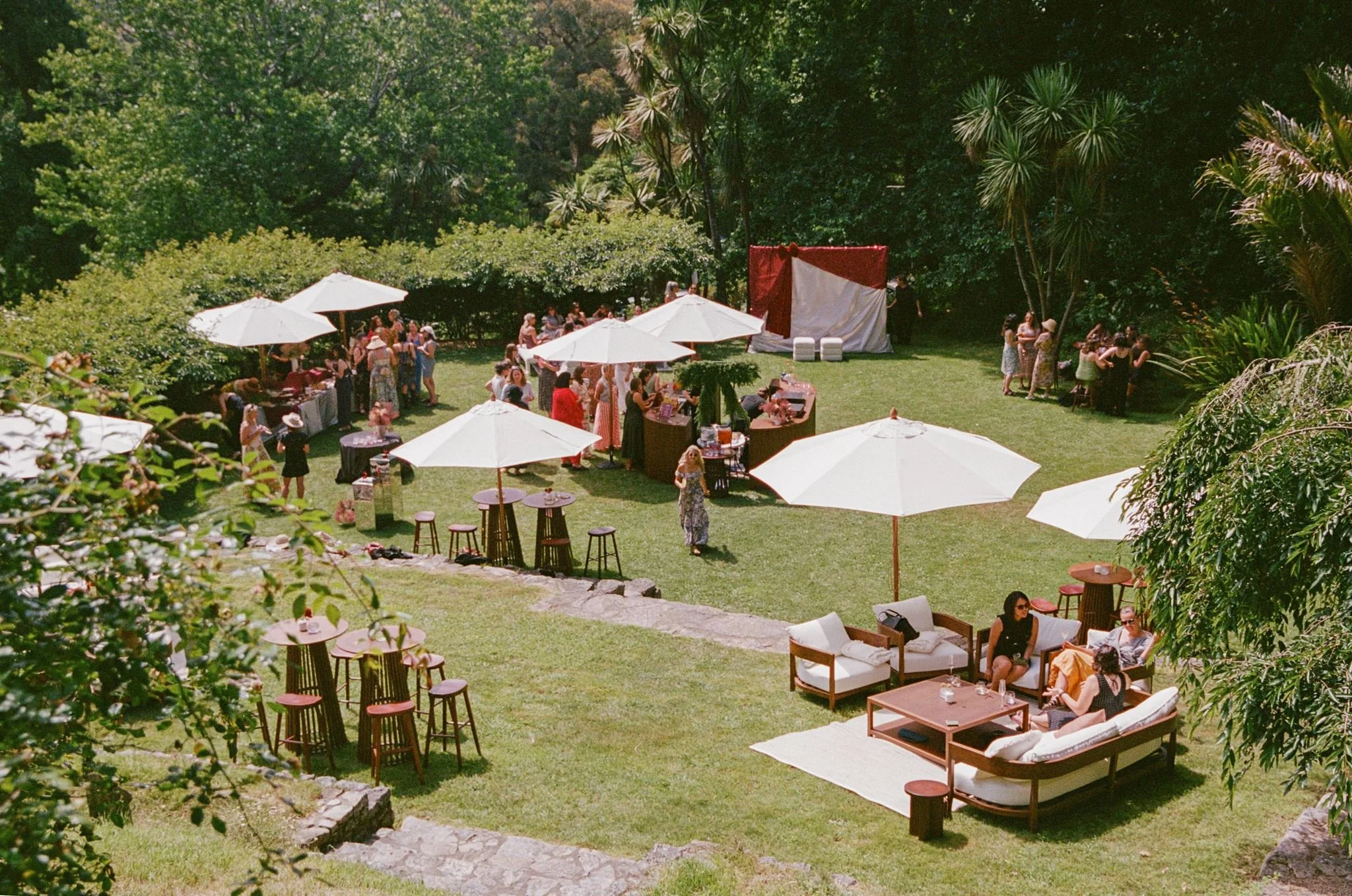 Outdoor gathering in a lush green garden with white umbrellas, people socializing, a small stage, and cozy seating areas.