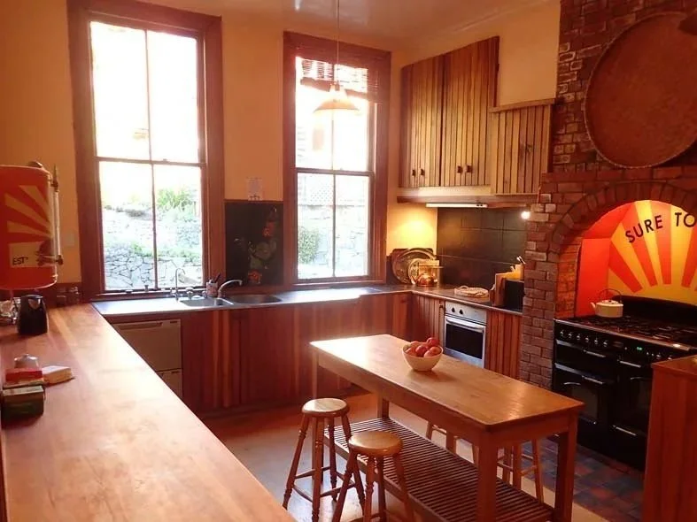 Sunlit kitchen with wooden cabinets, a small island with stools, a brick oven with a colorful sign that reads 'Sure to' in the background, and large windows letting in natural light.