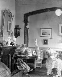 Black and white photo of a vintage living room with a fireplace, framed pictures on the wall, and chairs with patterned upholstery.
