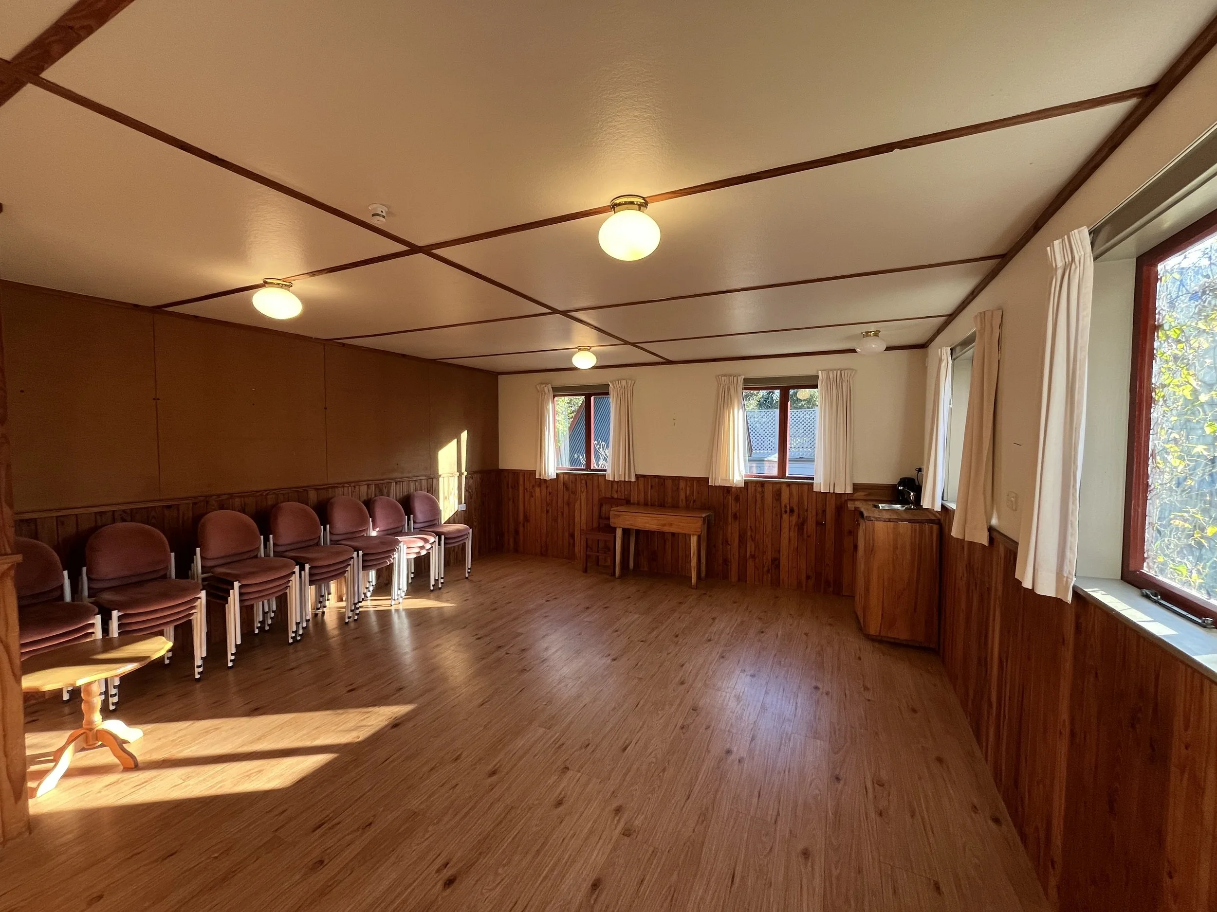 A spacious room with wooden flooring and wood-paneled walls, featuring multiple windows with white curtains, hanging light fixtures on the ceiling, and a row of chairs along one wall.