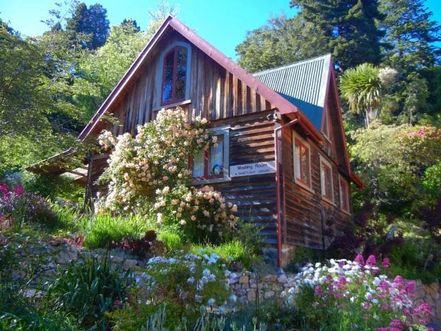 A rustic wooden house with a steep roof, surrounded by blooming flowers and greenery, under a clear blue sky.