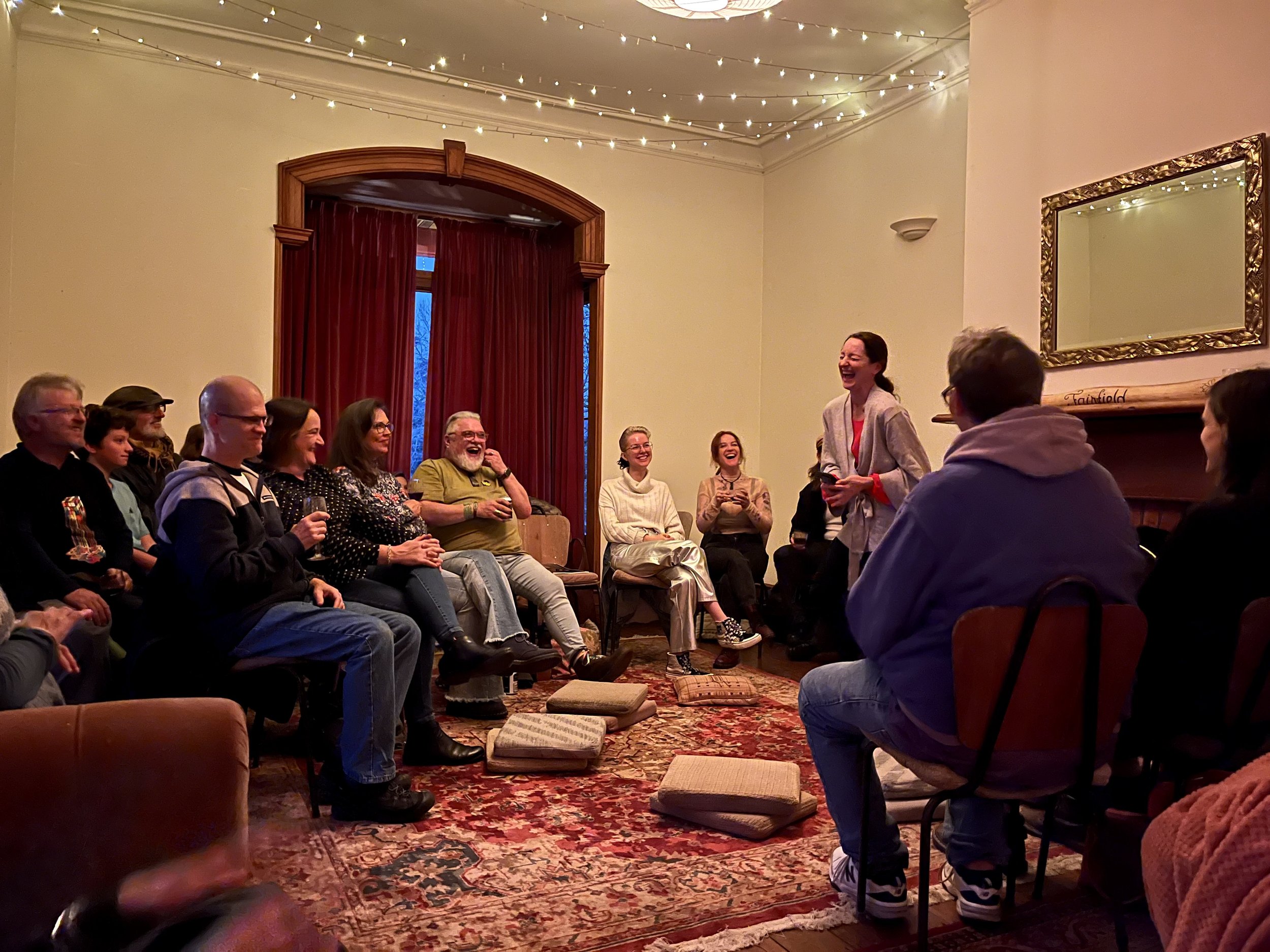 Group of people sitting in a semi-circle, laughing and smiling, during a gathering or event in a warmly lit room decorated with string lights, a large mirror, and a patterned rug. A woman stands speaking to the group.