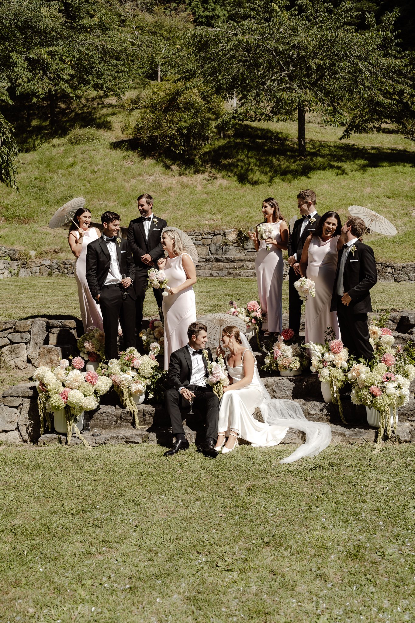 A wedding party posing outdoors on stone steps decorated with flowers, including brides and grooms, some holding umbrellas, and others holding bouquets, on a sunny day with a grassy yard and trees in the background.