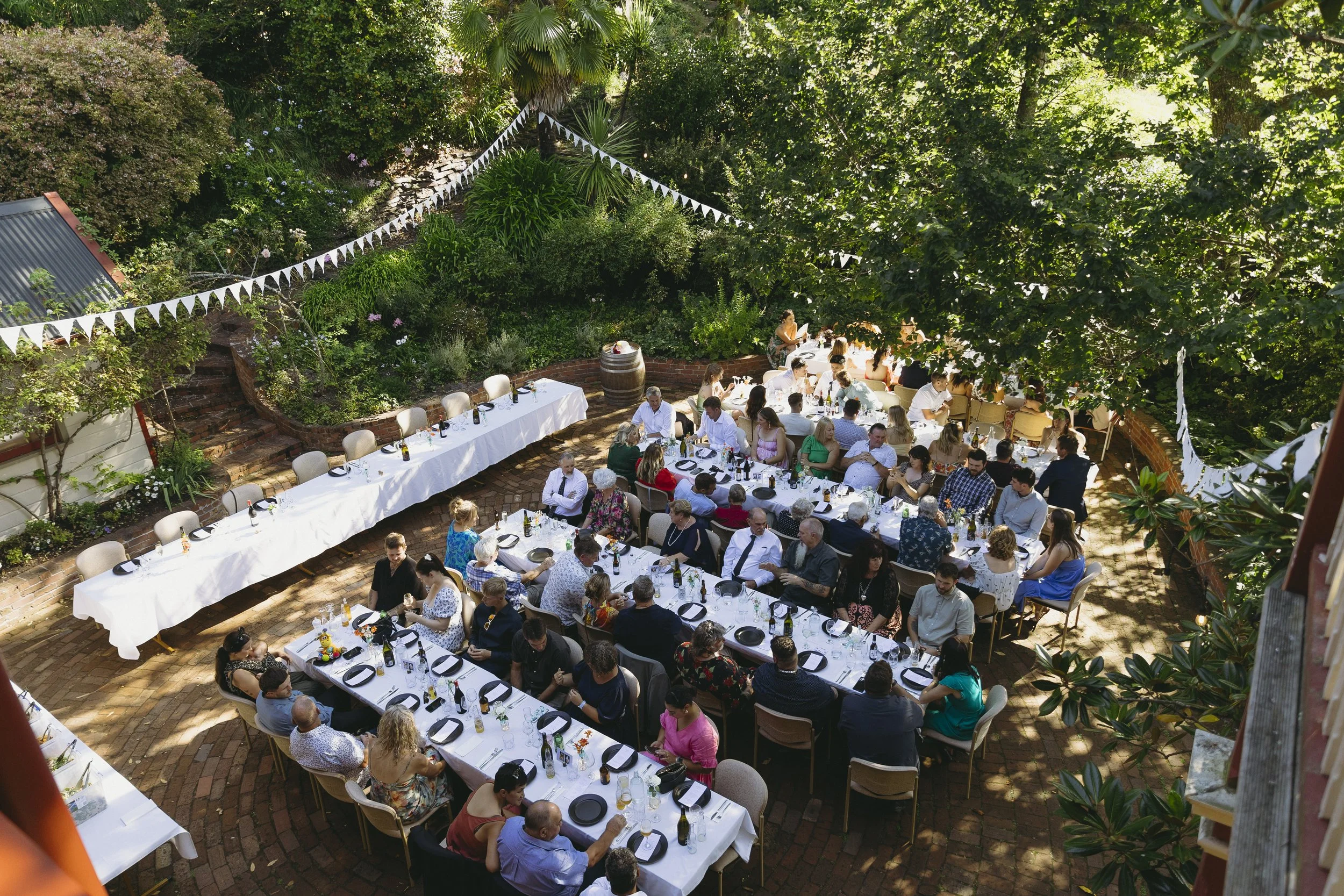 Outdoor gathering with multiple long tables covered with white tablecloths, set with black plates and drinks, surrounded by people, under a canopy of trees and string lights, in a lush garden setting.