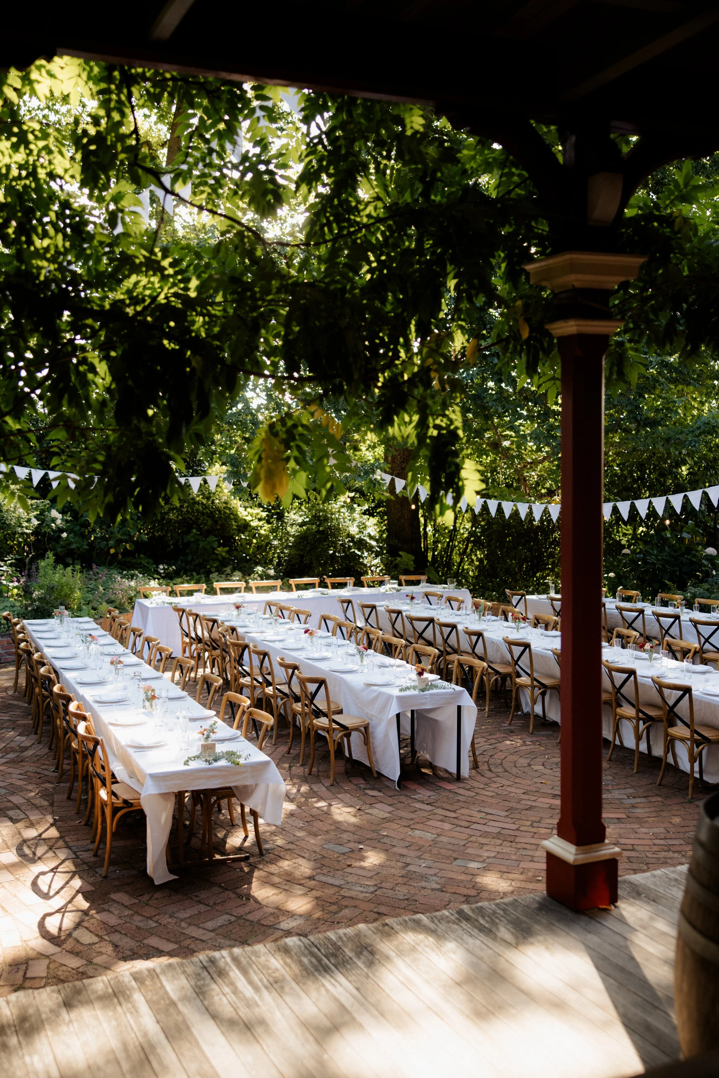 Outdoor event space with long tables covered in white tablecloths, decorated with small floral centerpieces, arranged for a gathering under a canopy of trees with hanging bunting banners.