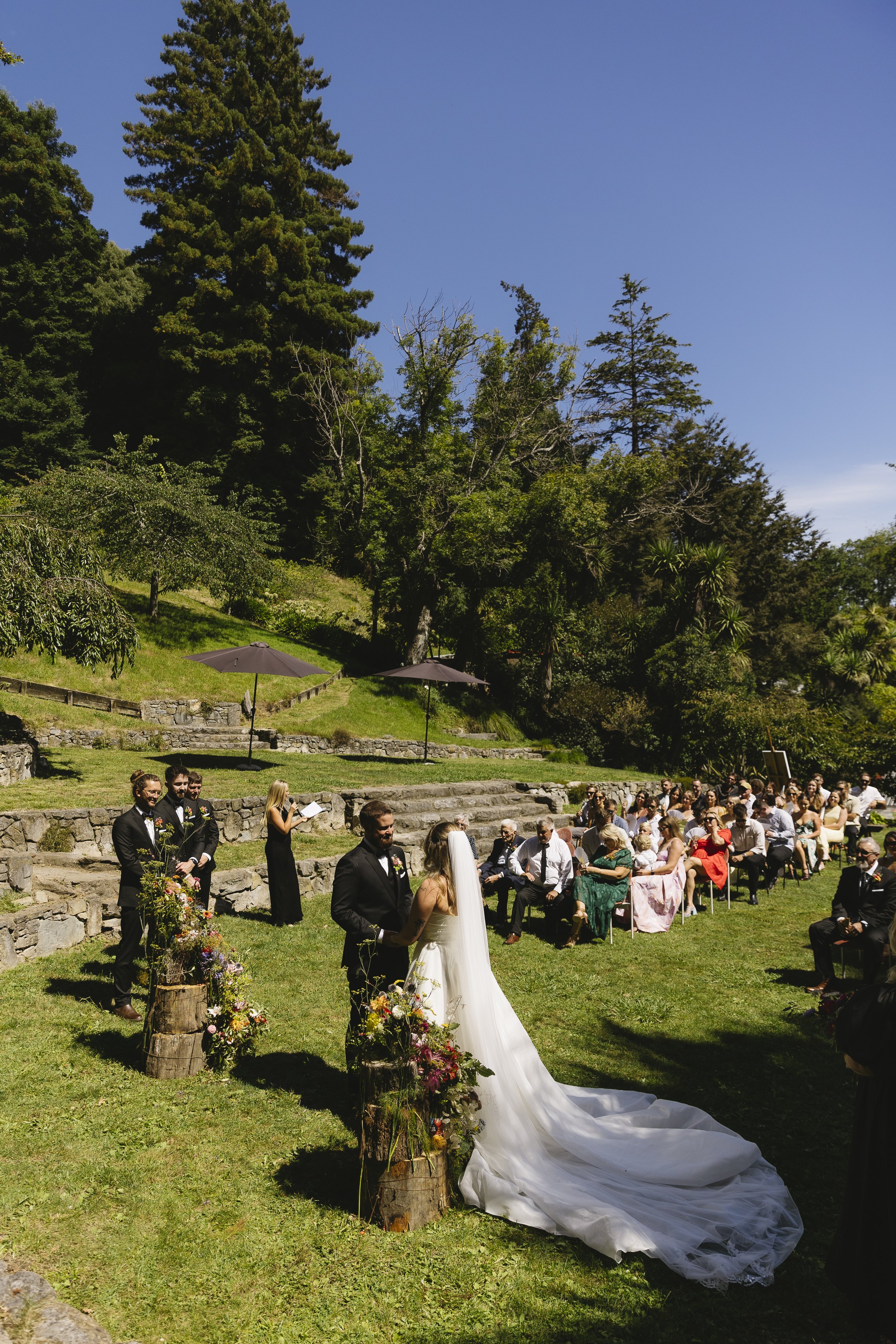 Outdoor wedding ceremony on grass with bride and groom, surrounded by seated guests, under a large tree and blue sky.
