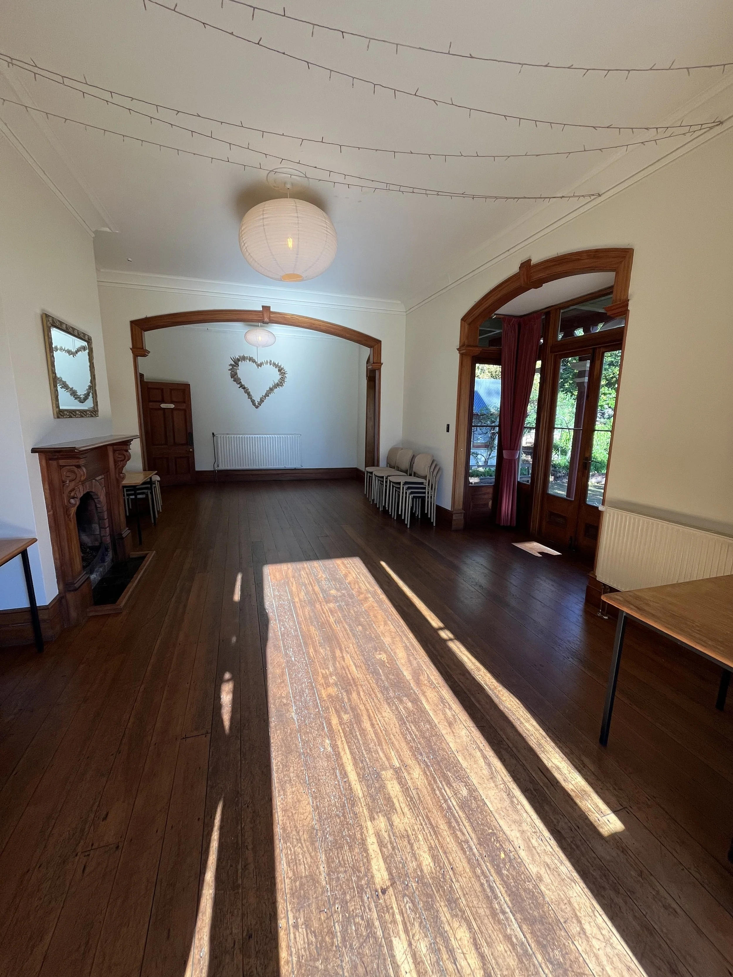 A room with a wooden floor, a white ceiling with hanging string lights, a paper lantern ceiling light, wooden door and window frames, a fireplace, and a small stack of chairs near the wall. Sunlight streams through the window and door, illuminating t