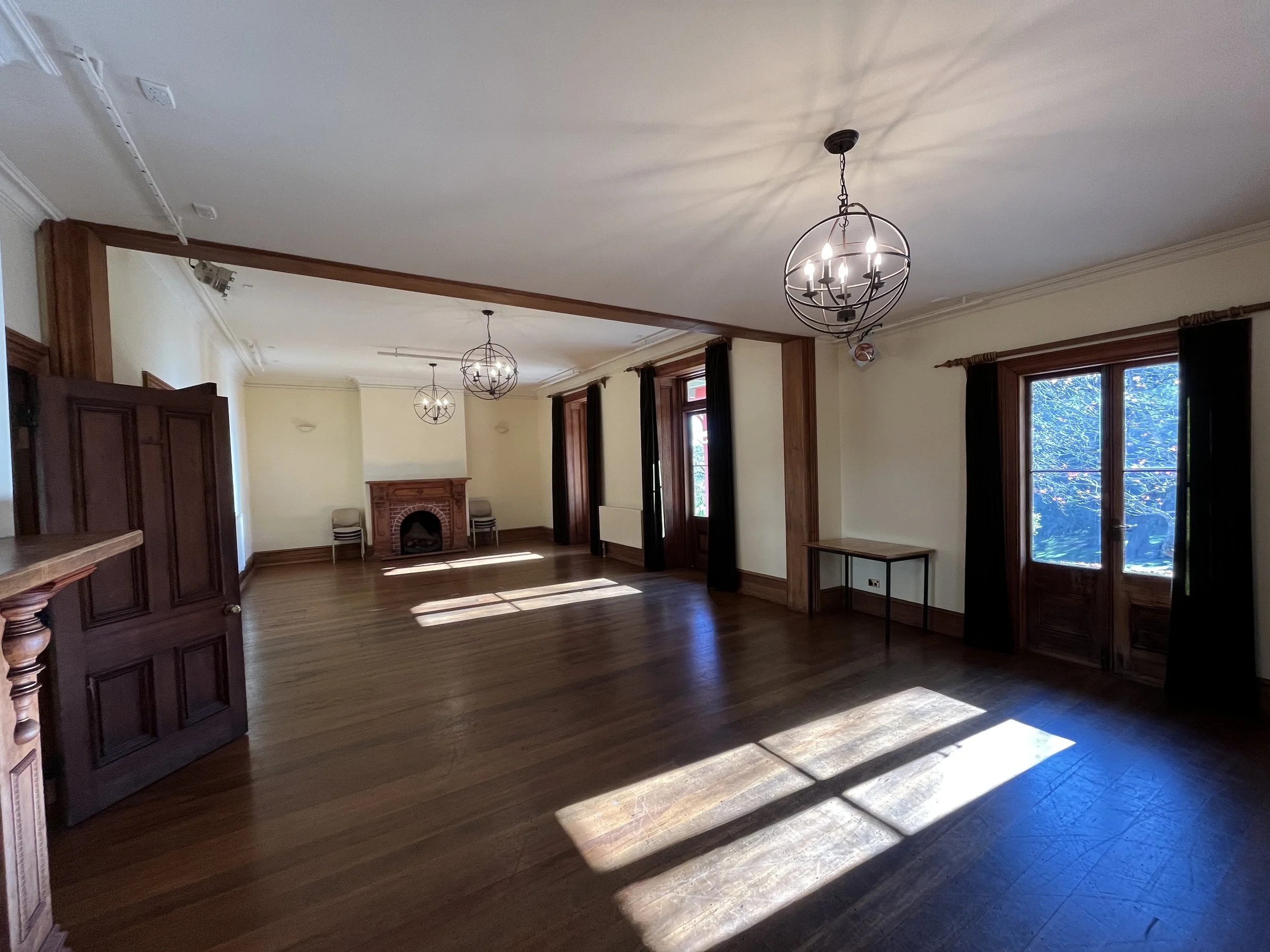 A spacious, well-lit room with wooden floors, three spherical chandeliers hanging from the ceiling, and large windows with outside greenery visible. The room features a brick fireplace and dark curtains.