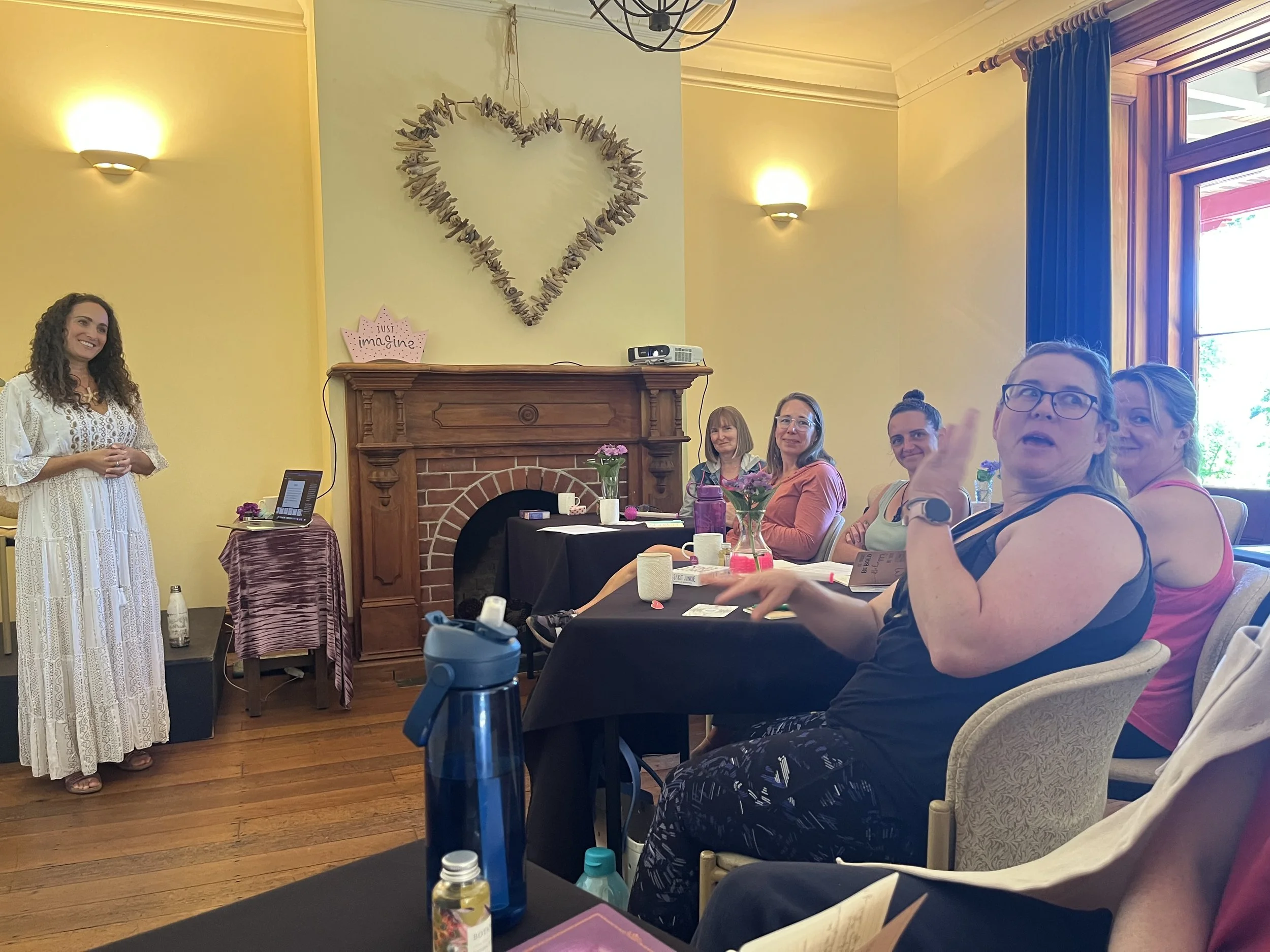 A group of women sitting at a table during a gathering or meeting in a room with a fireplace and a heart-shaped decoration made of driftwood on the wall. The women are engaged and smiling, with some looking towards the camera. There are flowers, wate